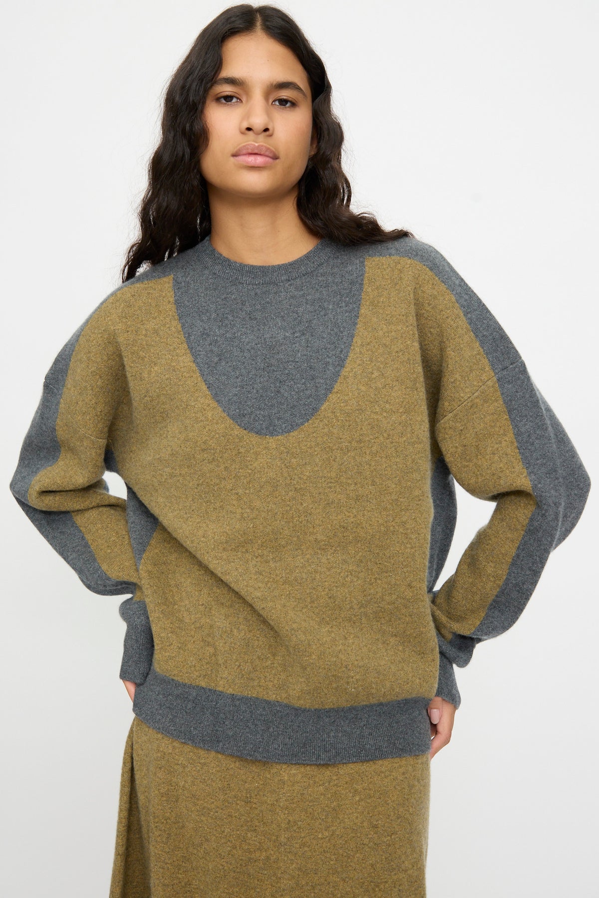A person with long dark hair is wearing the Extreme Cashmere No. 518 Trompe Sweater in Hunter and Felt, a loose-fitting color-blocked knit with a classic crew neckline, standing against a plain white background.