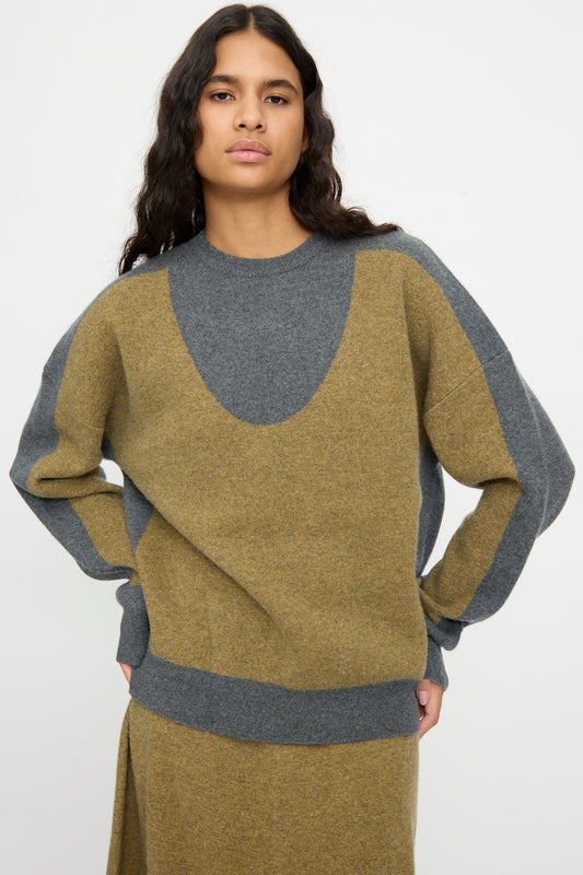 A person with long dark hair is wearing the Extreme Cashmere No. 518 Trompe Sweater in Hunter and Felt, a loose-fitting color-blocked knit with a classic crew neckline, standing against a plain white background.