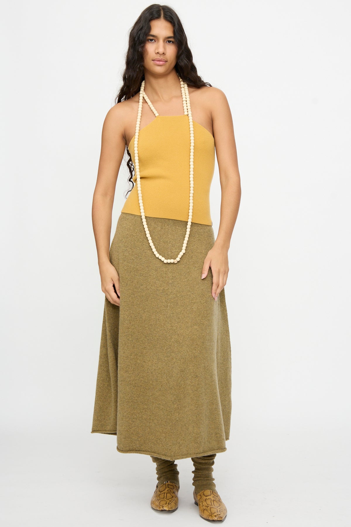A woman with long dark hair wears the No. 519 Candy Top in Wheat by Extreme Cashmere—a cashmere blend strapless top—paired with an olive skirt, a long beaded necklace, leg warmers, and brown shoes, standing against a plain white background.