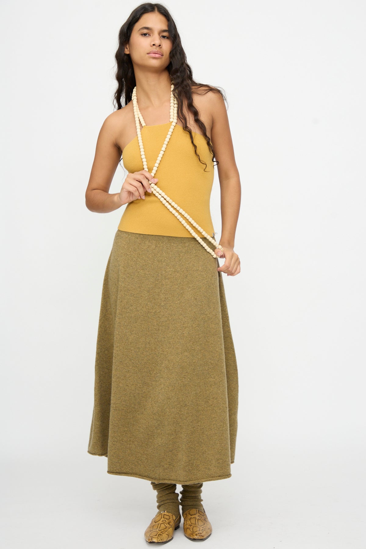 Wearing the Extreme Cashmere No. 519 Candy Top in Wheat, paired with an olive green midi skirt, tan sandals with socks, and layered bead necklaces, this individual stands against a plain white background.