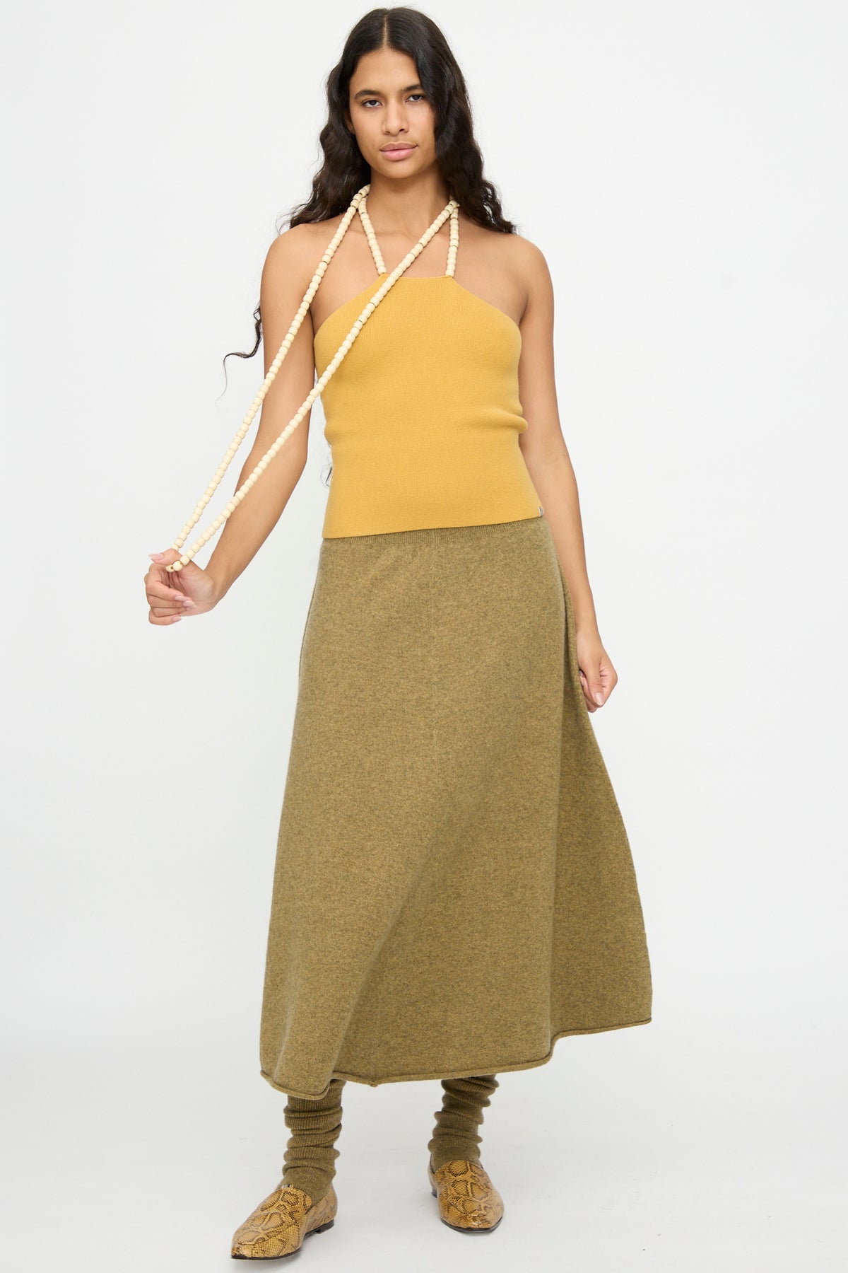 A woman with long dark hair wears the Extreme Cashmere No. 519 Candy Top in Wheat, styled with an olive maxi skirt, matching leg warmers, and beige shoes, standing against a plain white background.