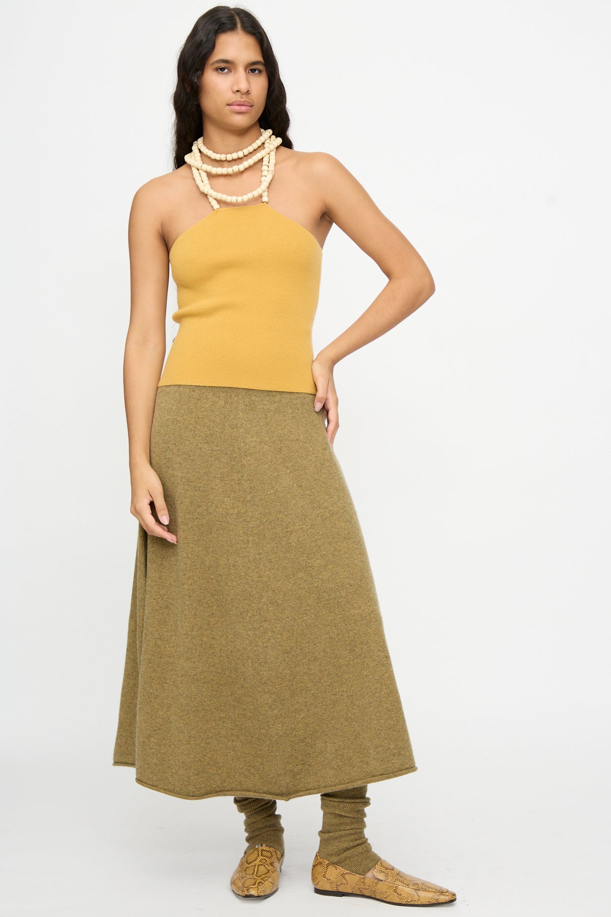 A woman dressed in the Extreme Cashmere No. 519 Candy Top in Wheat, paired with an olive green skirt, matching leg warmers, snake-print shoes, and layered pearl necklaces stands against a plain white background.