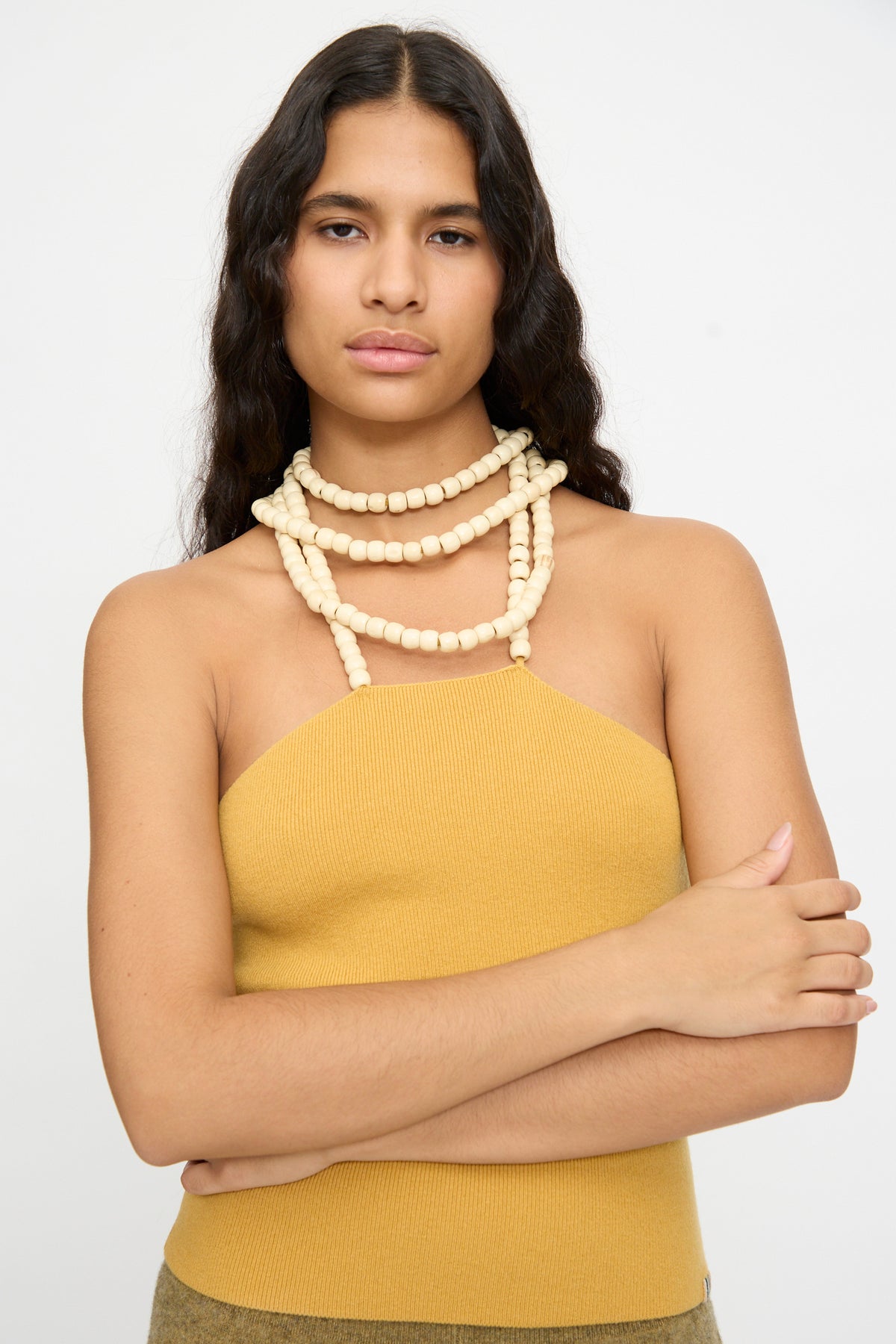 A woman with long dark hair wears the Extreme Cashmere No. 519 Candy Top in Wheat—a mustard yellow rib-knit halter—paired with a chunky cream bead necklace, standing with arms crossed against a plain light background.