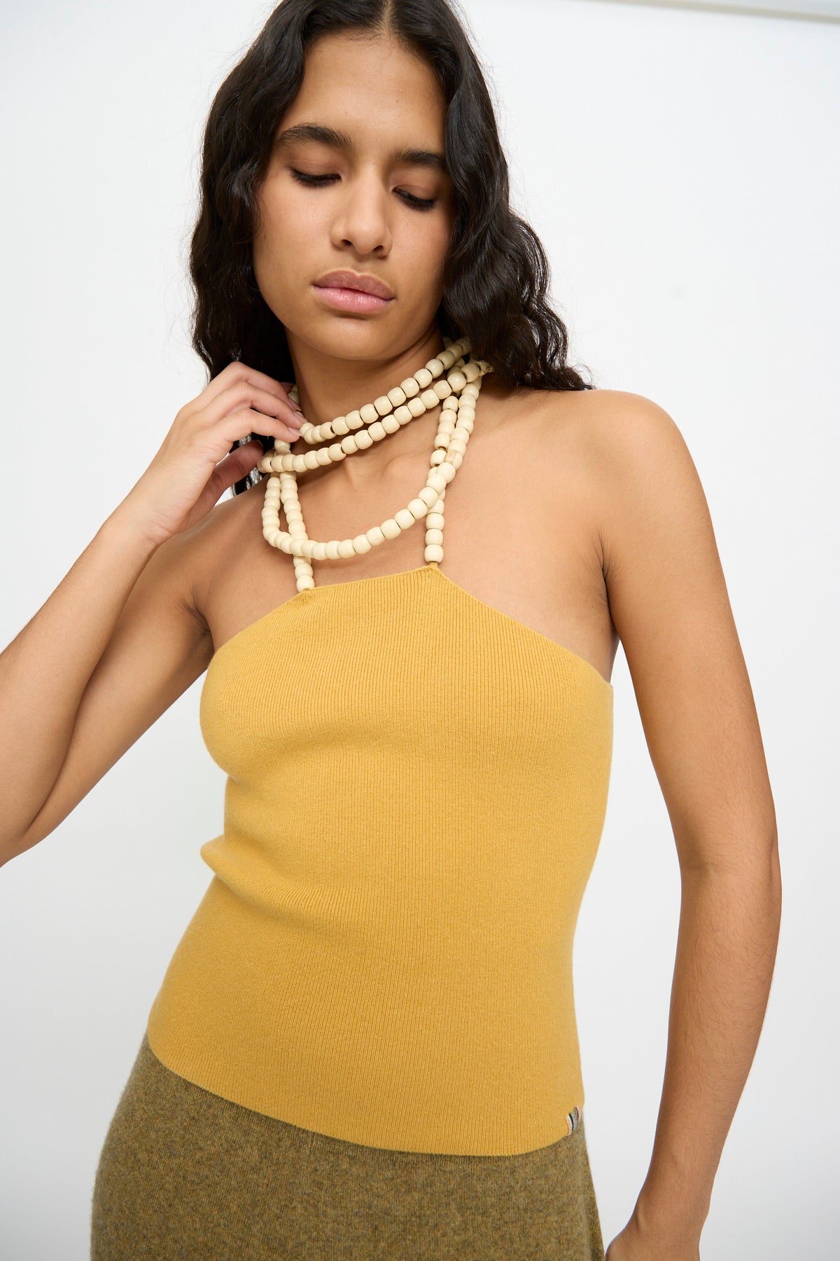 A person wearing the Extreme Cashmere No. 519 Candy Top in Wheat and layered beaded necklaces stands against a plain white background.