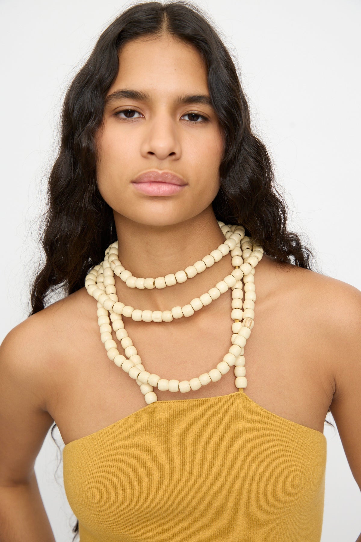 A woman with long wavy hair wears the Extreme Cashmere No. 519 Candy Top in Wheat and layered chunky cream beaded necklaces, standing against a plain white background.