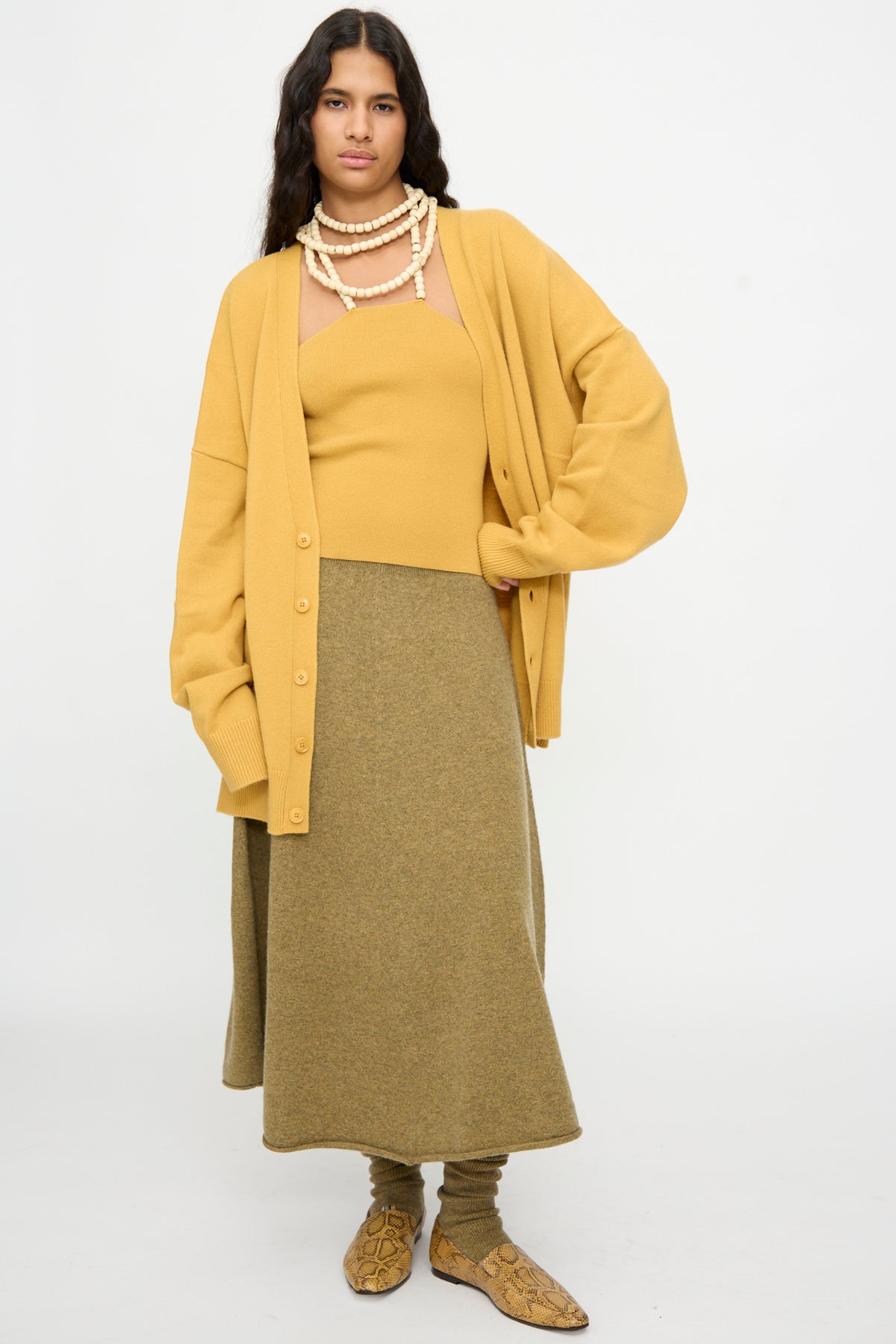 Woman wearing a mustard cardigan, an Extreme Cashmere No. 519 Candy Top in Wheat, olive skirt, layered necklaces, and snakeskin shoes stands against a plain white background.