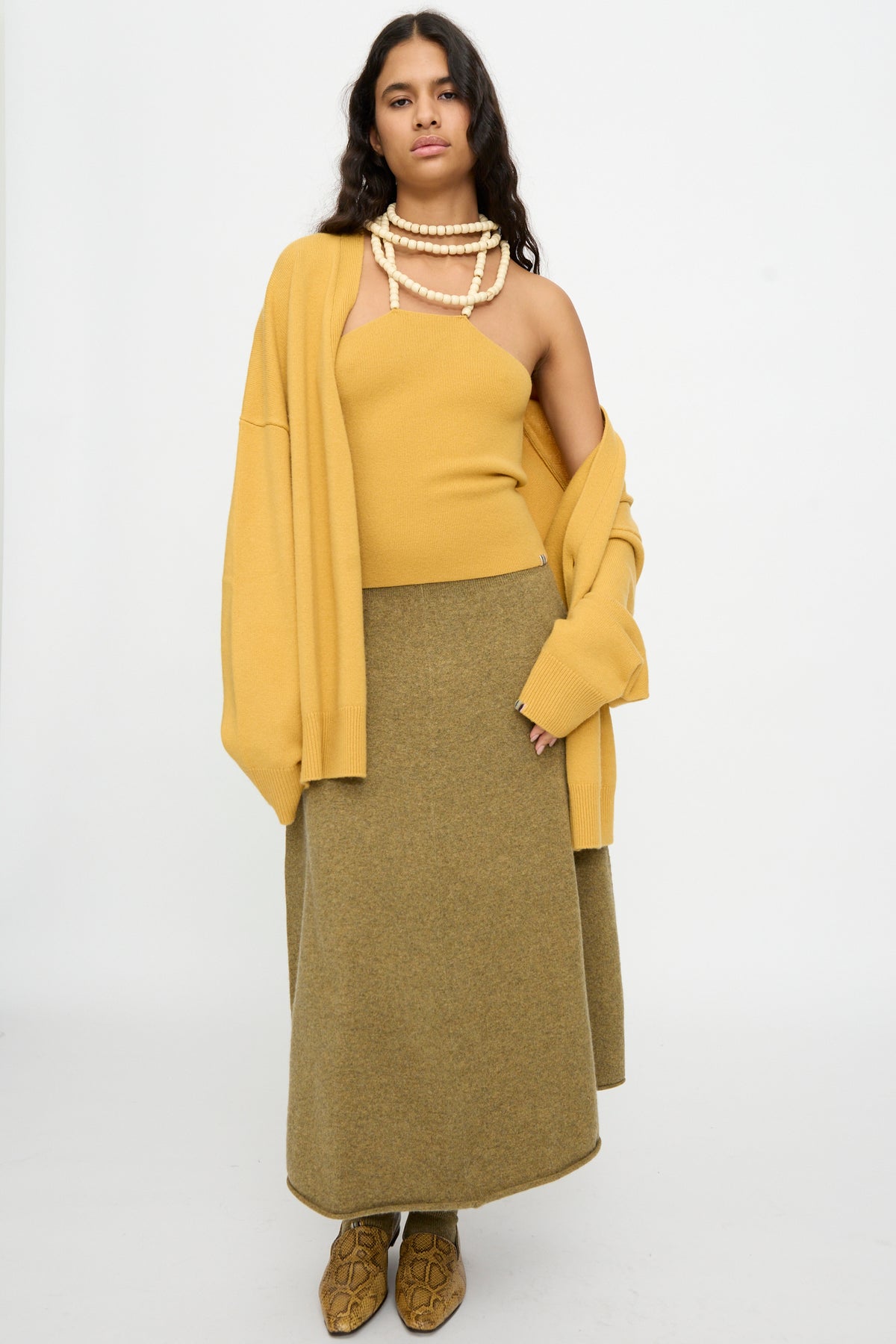 A woman stands against a plain background wearing the No. 519 Candy Top in Wheat by Extreme Cashmere, an oversized sweater, a long olive skirt, tan boots, and layered necklaces.