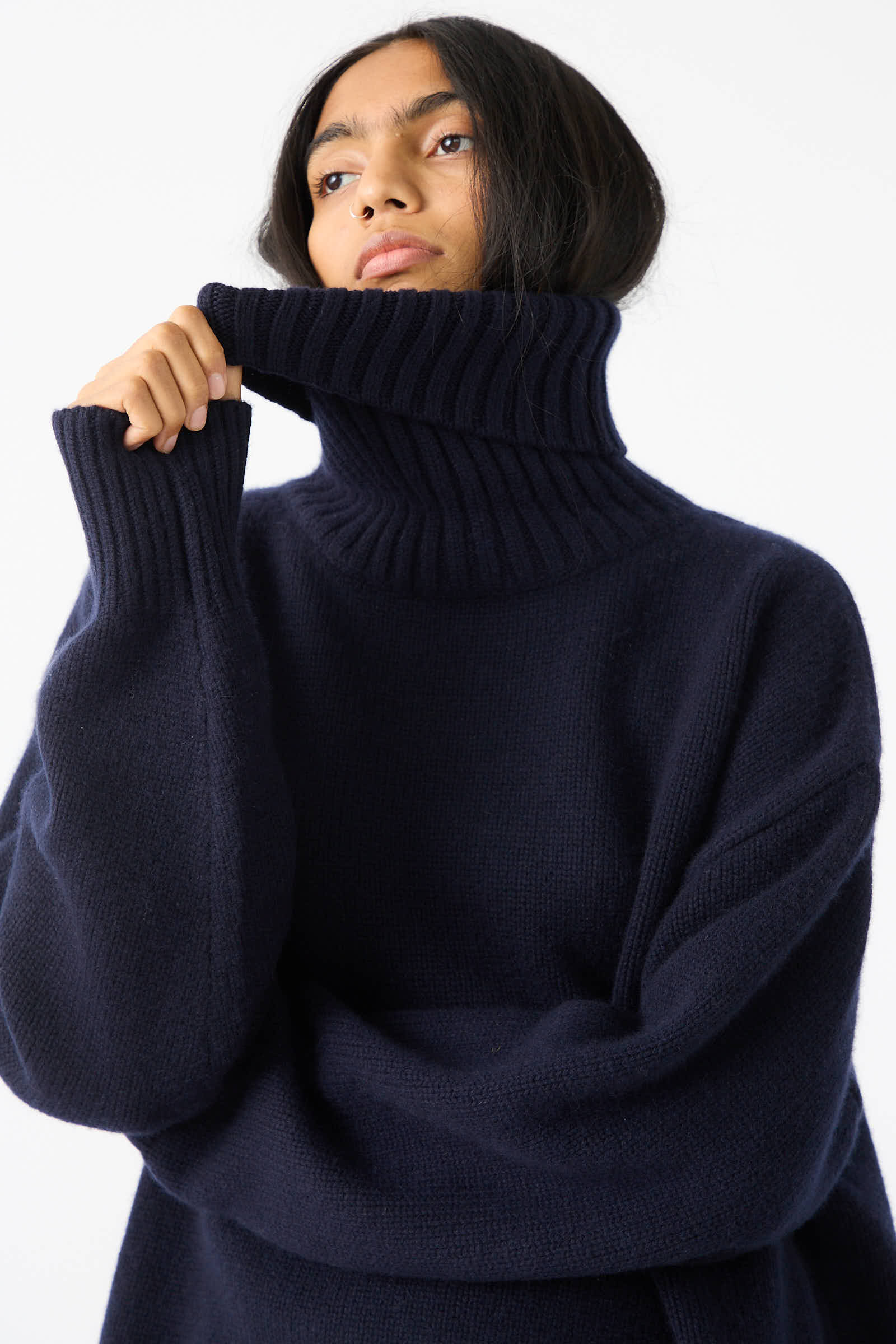 A person with long dark hair wears the No. 20 Oversize Extra Turtleneck Sweater in Navy by Extreme Cashmere, partially covering their mouth with their hand.