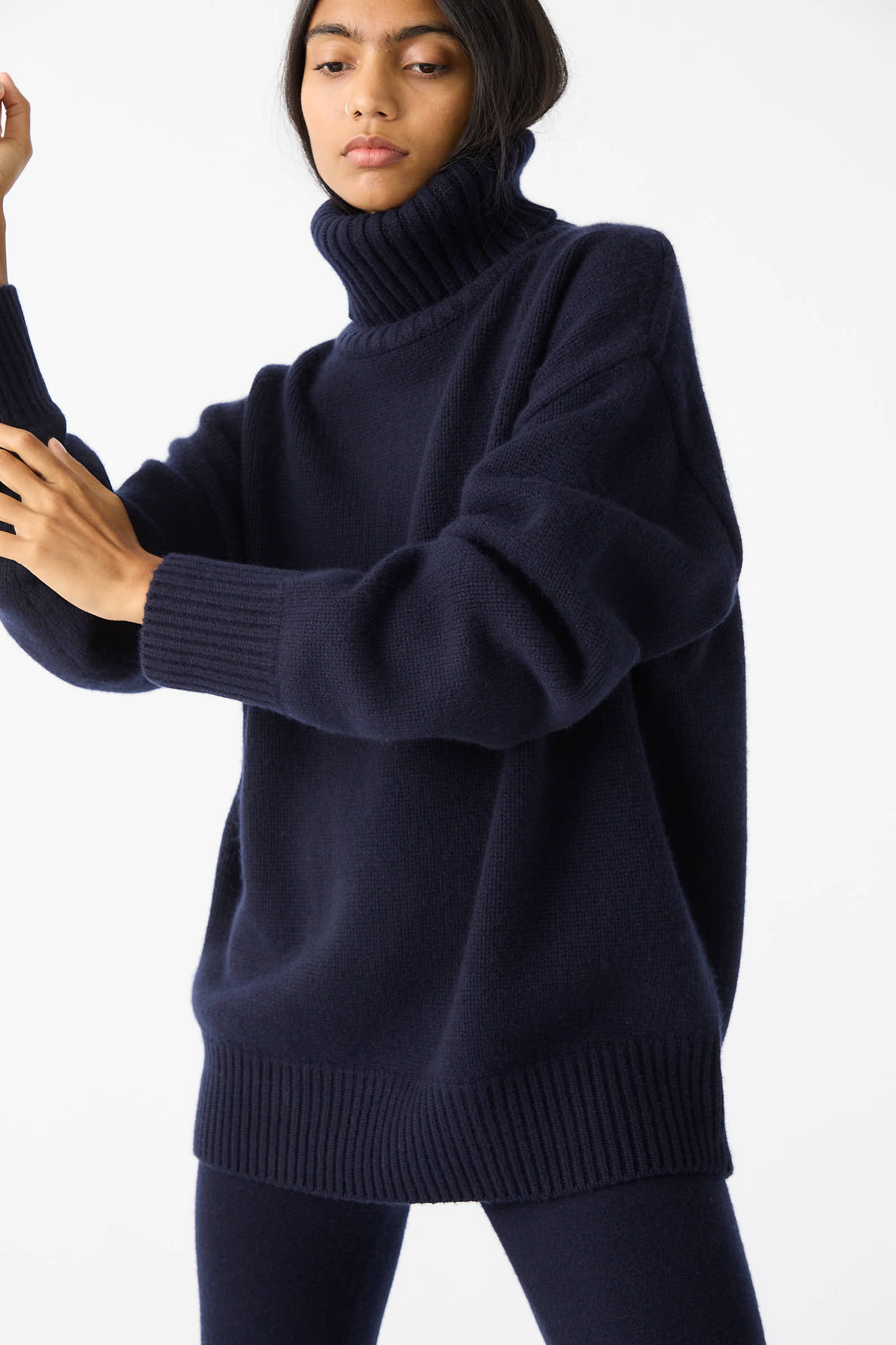 A person wearing an Extreme Cashmere No. 20 Oversize Extra Turtleneck Sweater in Navy and matching pants stands against a plain white background.