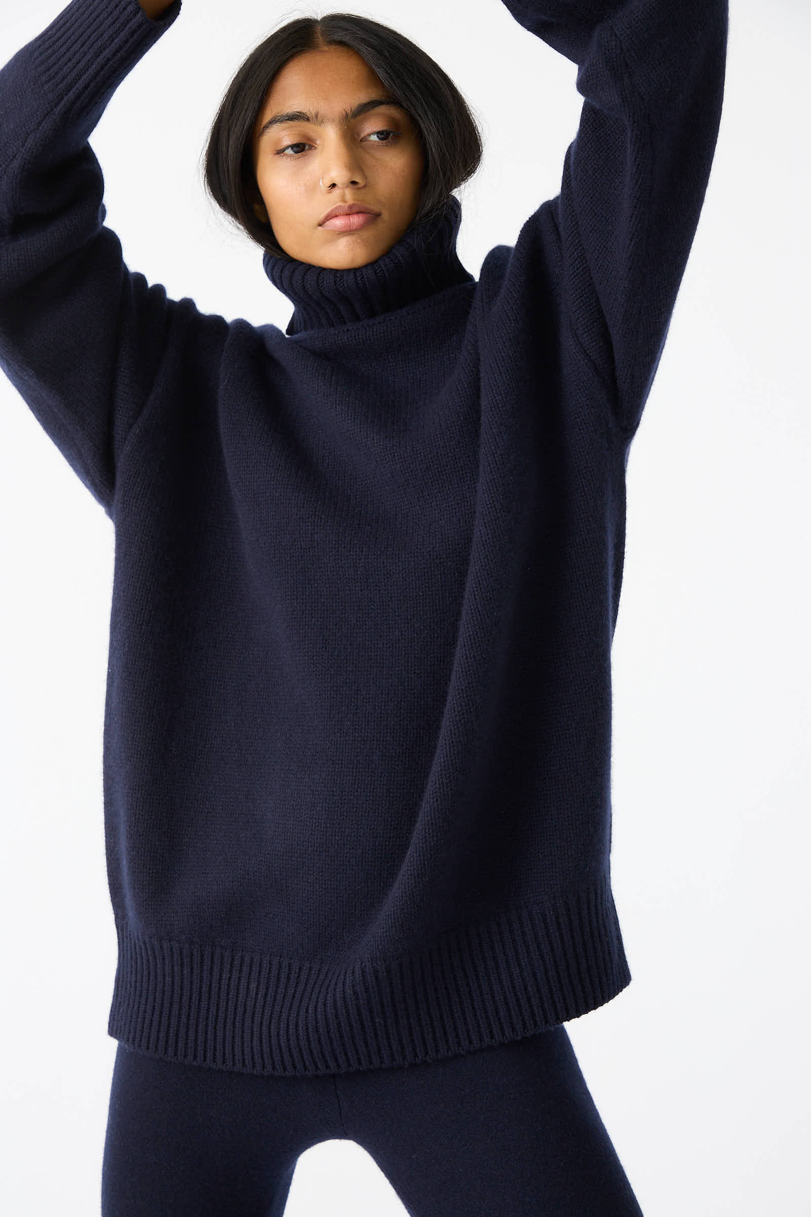 A person stands with raised arms, wearing the Navy No. 20 Oversize Extra Turtleneck Sweater from Extreme Cashmere and matching leggings, set against a plain white background, highlighting the cozy elegance of this oversized sweater.