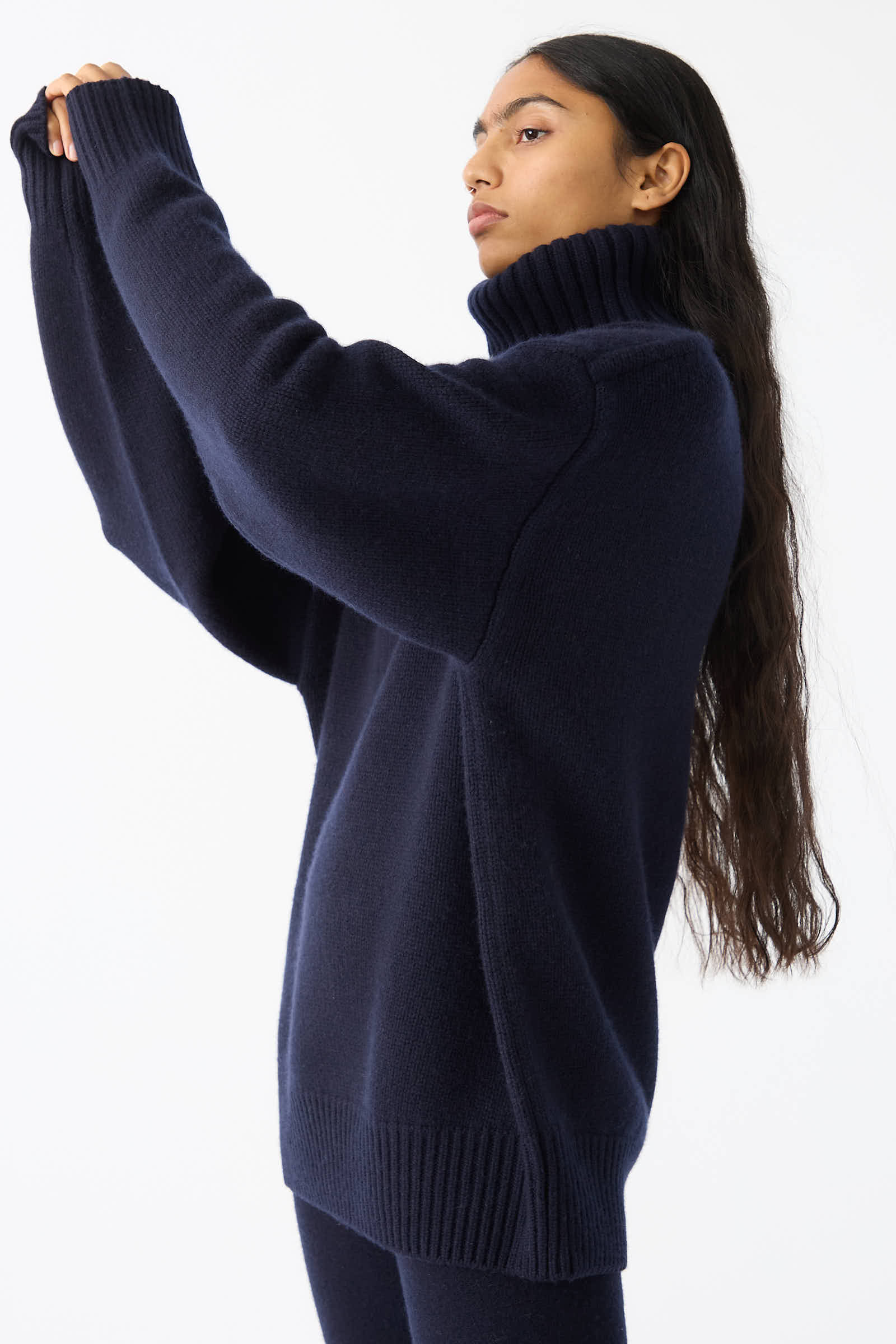 Individual with long hair in the No. 20 Oversize Extra Turtleneck Sweater by Extreme Cashmere in Navy, adjusting the sleeve with one arm raised. White background.