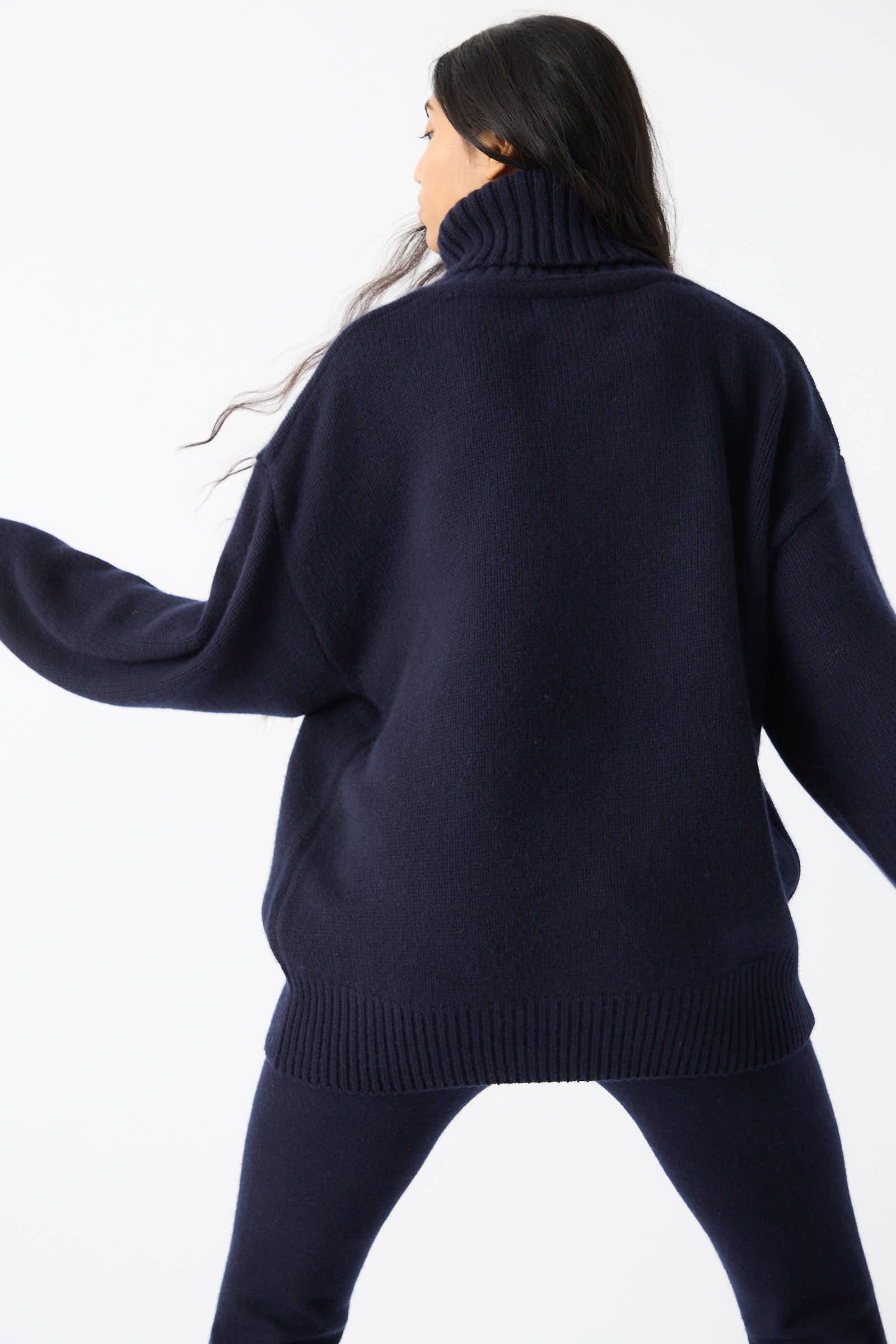 A person with long dark hair is wearing an oversized, navy No. 20 Extra Turtleneck Sweater by Extreme Cashmere, complemented by matching pants, standing with their back to the camera.