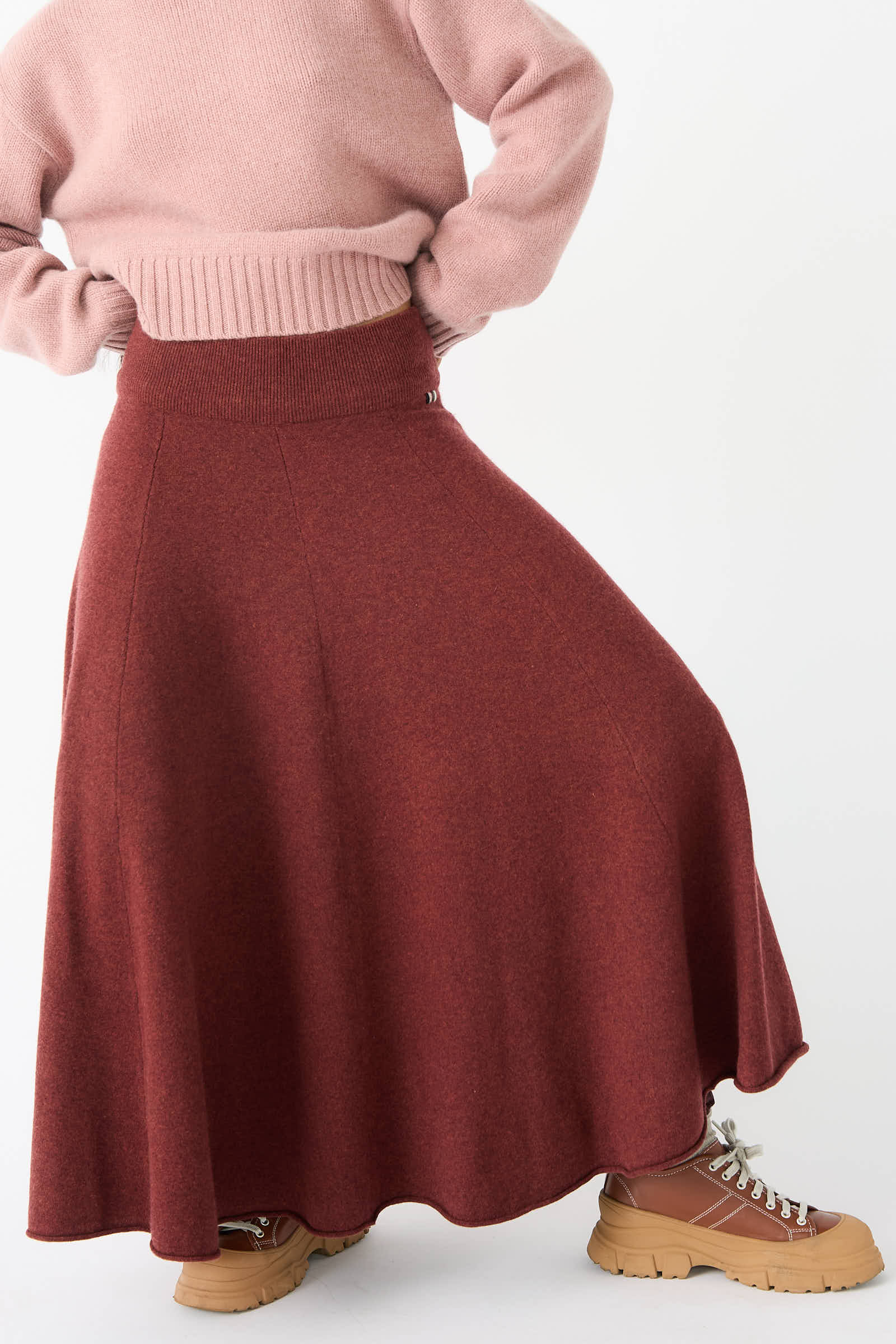 Person wearing a pink sweater, Extreme Cashmere's No. 313 Twirl Skirt in Maroon with an adjustable waist, and light brown chunky boots.