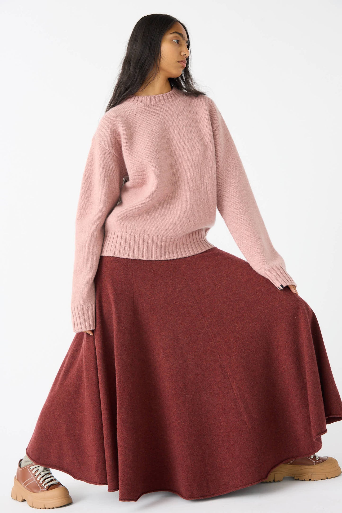 A person wearing a pink sweater and Extreme Cashmere's No. 313 Twirl Skirt in Maroon poses against a plain white background. They are also wearing tan platform shoes.