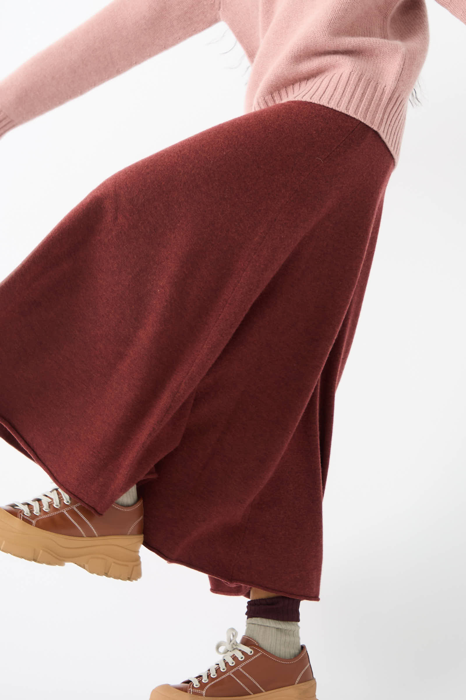 A person wearing a pink sweater, Extreme Cashmere's No. 313 Twirl Skirt in Maroon with an adjustable waist, brown platform sneakers, and beige socks is captured mid-movement on a white background.