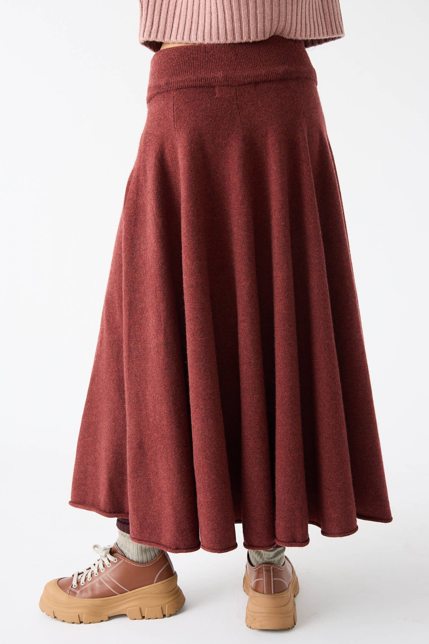 A person wearing an Extreme Cashmere No. 313 Twirl Skirt in Maroon, a pink sweater, gray socks, and tan shoes with thick soles.