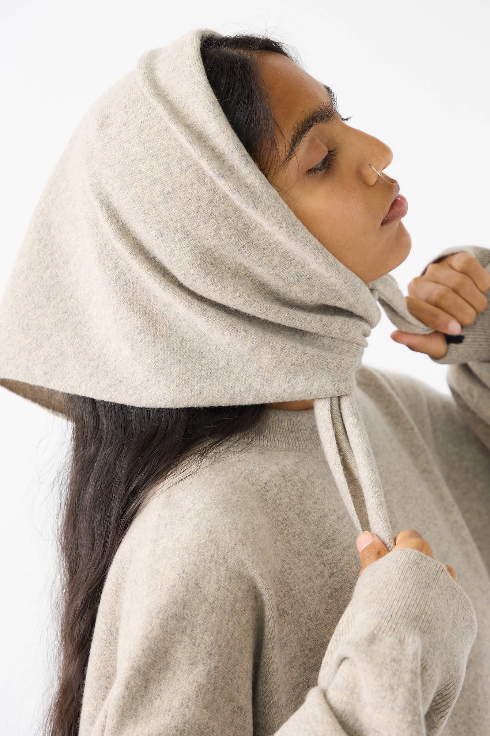 A person wearing a beige No. 35 Bandana in Moss from Extreme Cashmere adjusts the drawstrings of the hooded sweater. The cashmere blend fabric looks incredibly soft. The person is shown from the side against a plain white background.