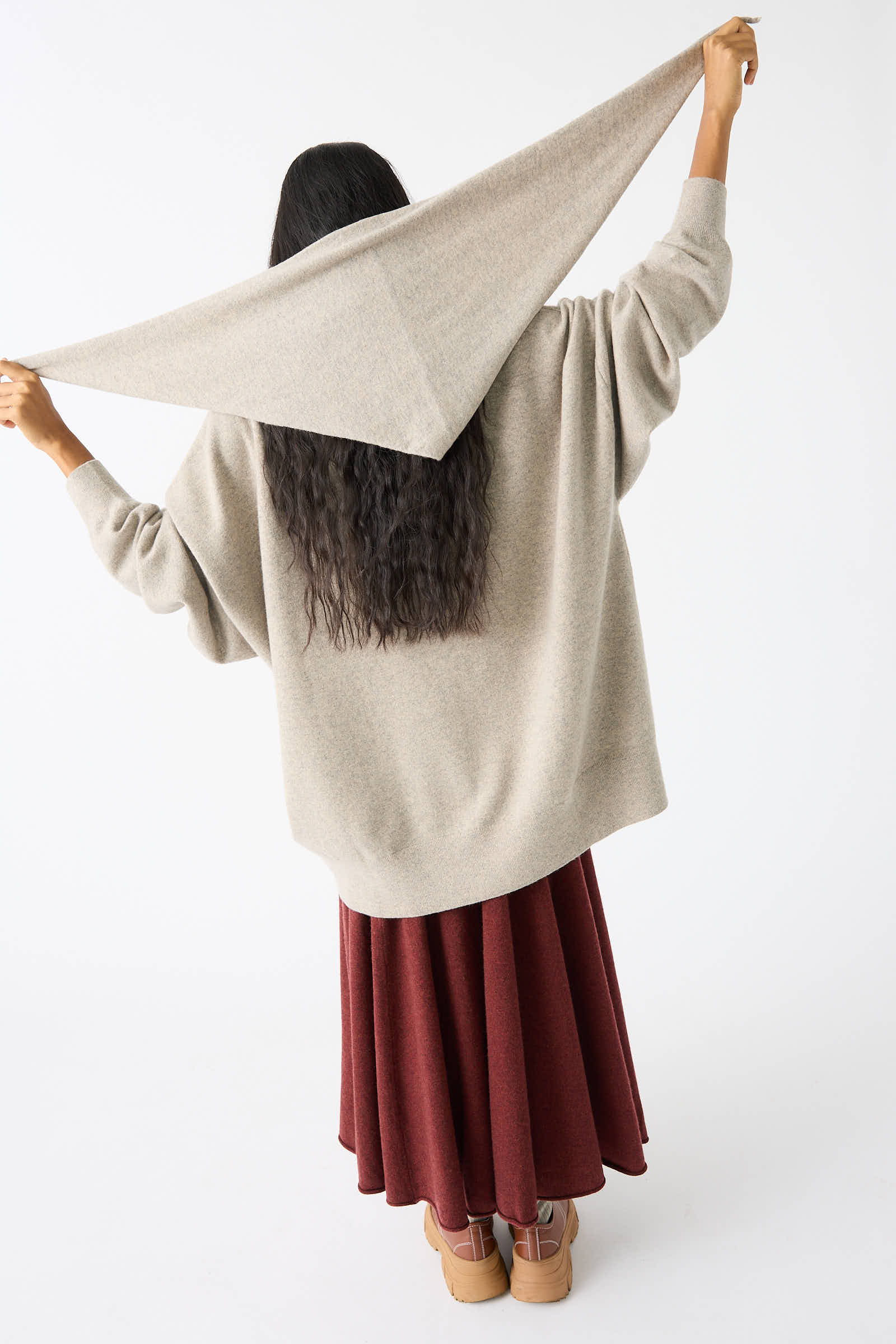 A person with long hair is seen from behind, wearing a beige sweater, red skirt, and tan boots, holding out the No. 35 Bandana in Moss by Extreme Cashmere up behind their head.