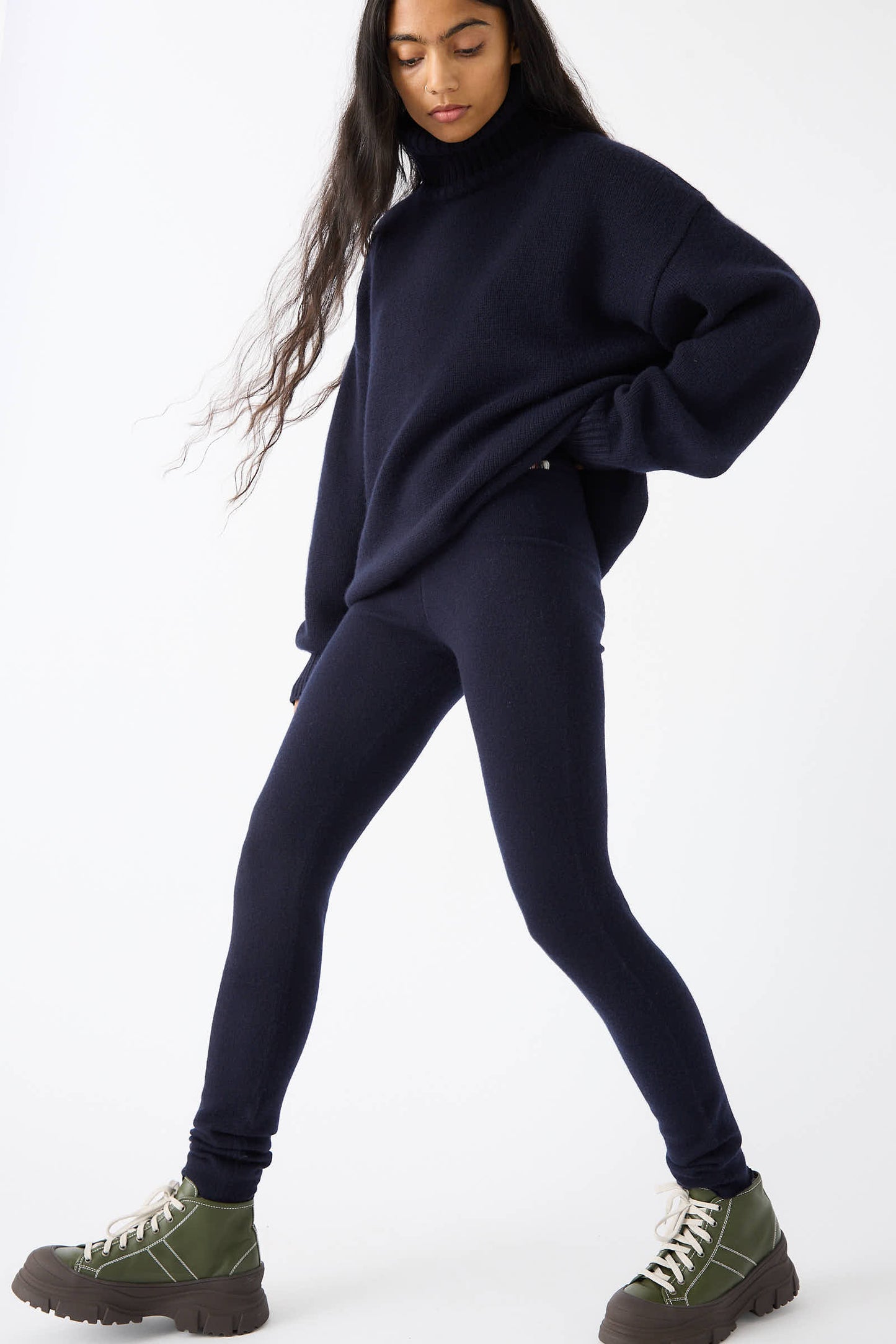 A person with long hair wearing a dark oversized sweater, Extreme Cashmere's No. 372 Beat Trouser in Navy, and green lace-up boots poses against a plain white background.