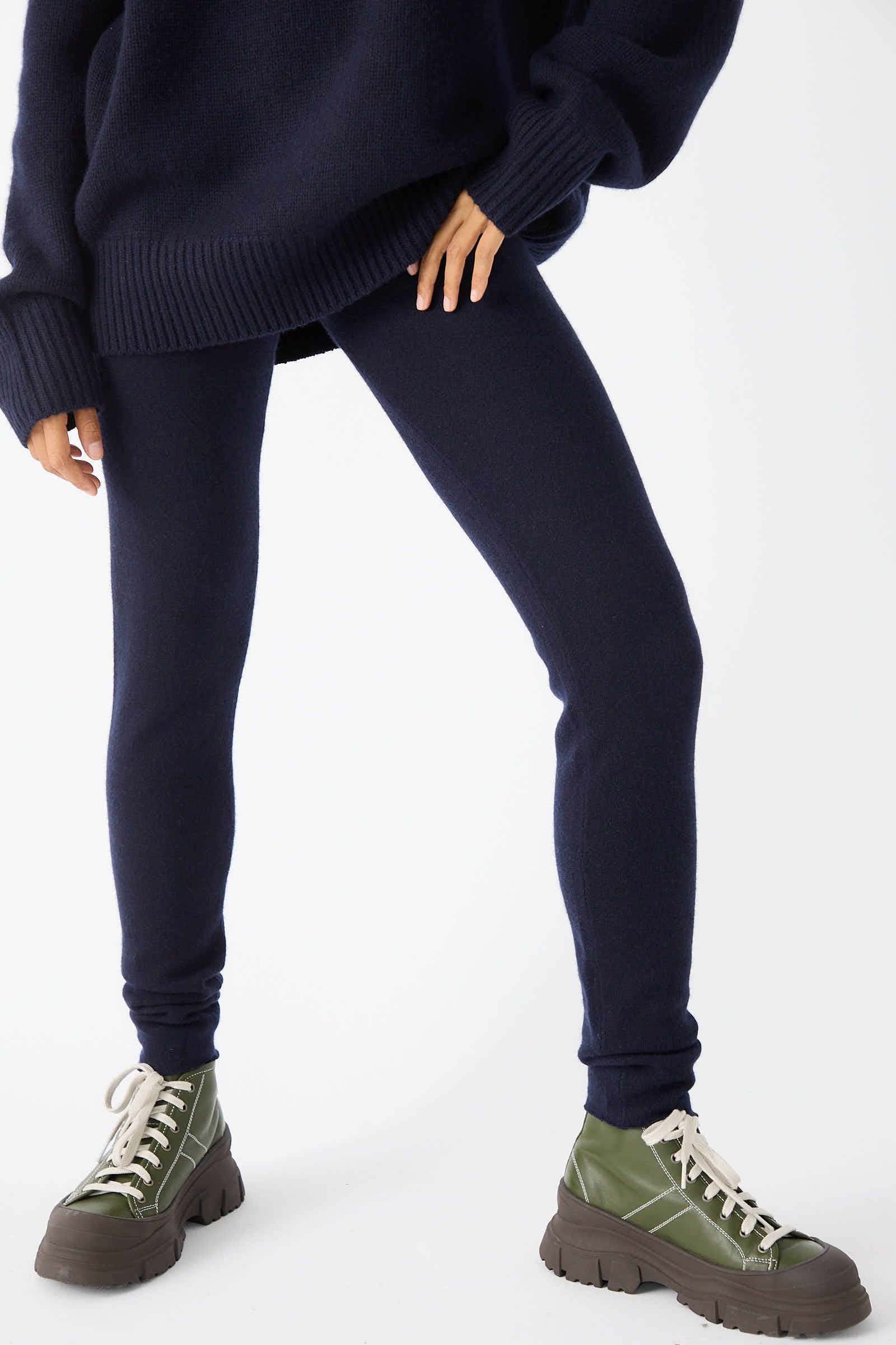 A person wearing a navy blue Extreme Cashmere No. 372 Beat Trouser with a rib-knit elasticated waist, and green lace-up boots against a white background.