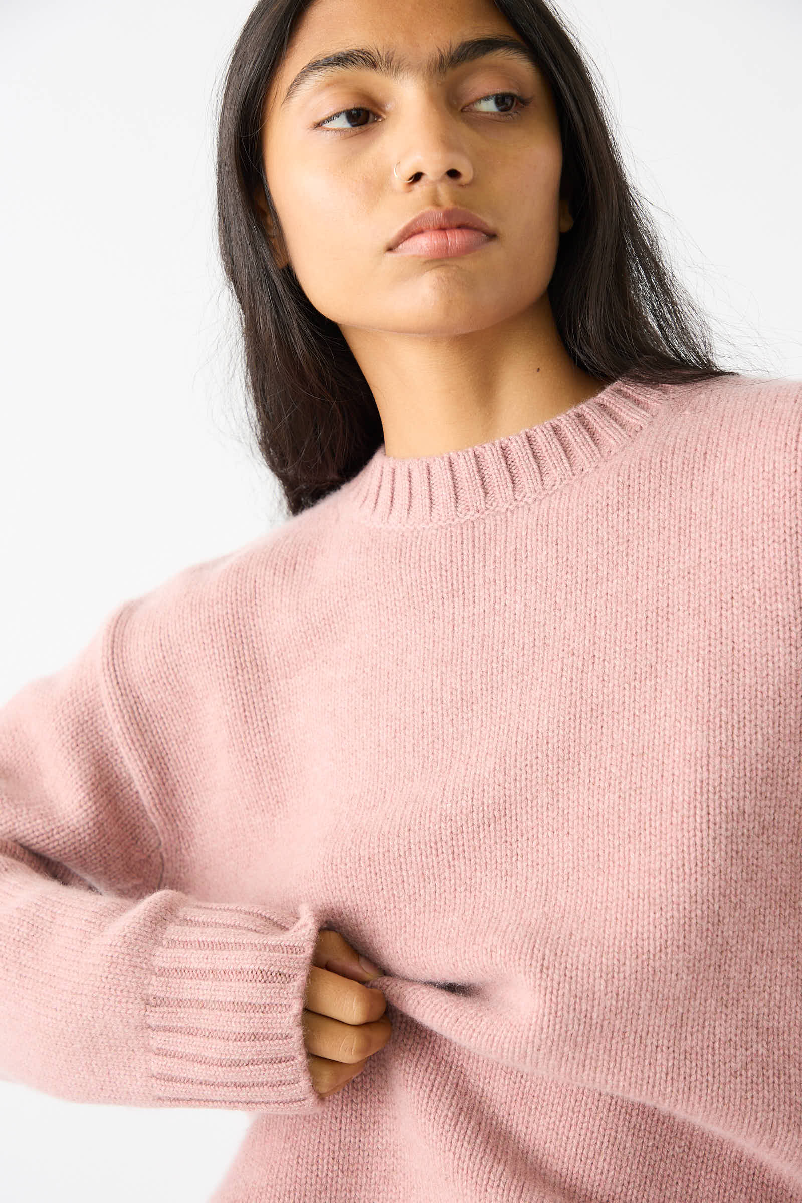 A person with long dark hair wearing the No. 373 Oldie Sweater in Moor by Extreme Cashmere looks to the side against a plain background.