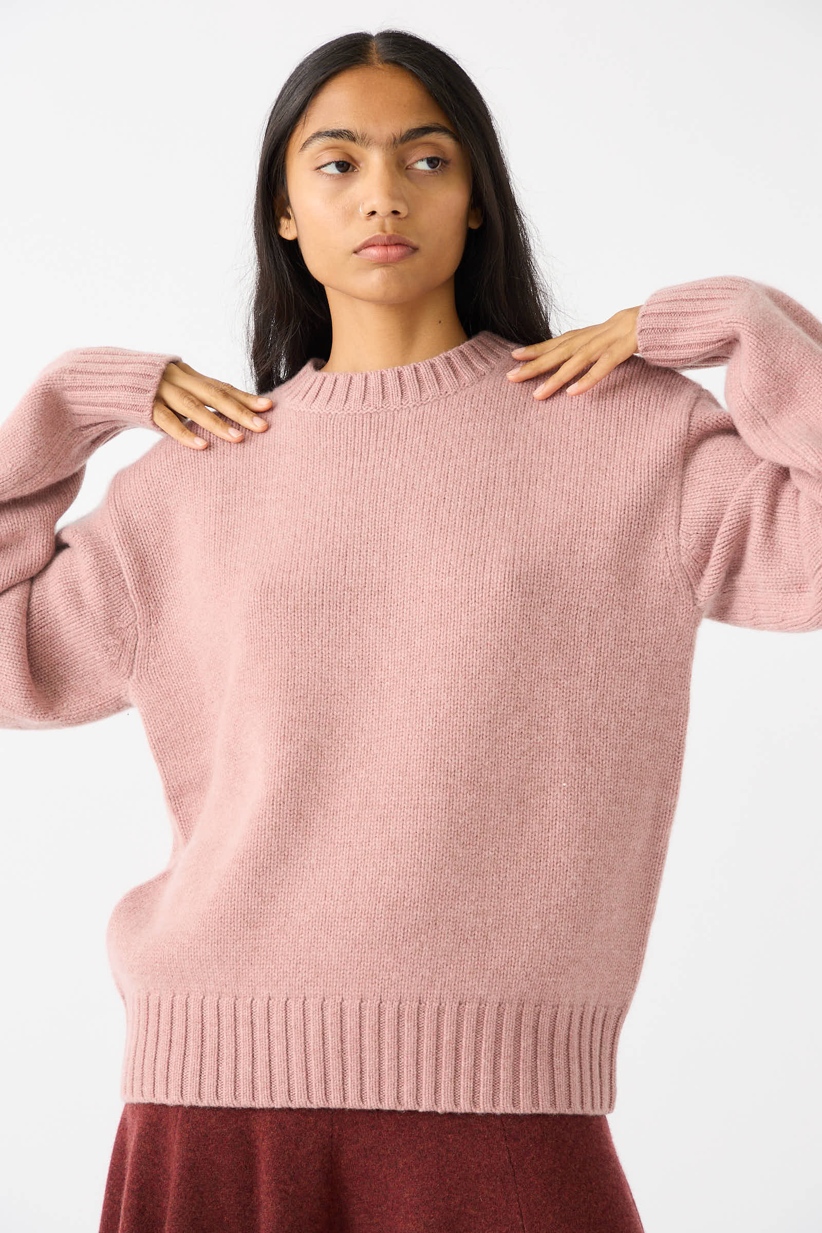 A person with long hair wears the No. 373 Oldie Sweater in Moor by Extreme Cashmere, a pink, one-size, heavy-knit cashmere crew neck sweater, adjusting the sleeves while looking slightly to the side.