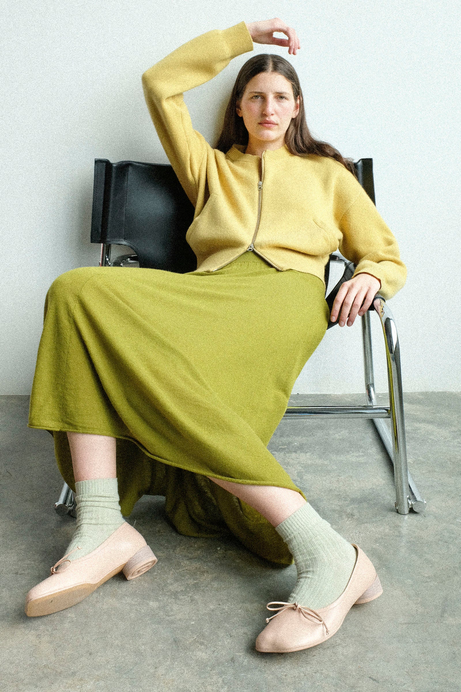 Woman sitting on a chair wearing Extreme Cashmere's No. 478 Xtra Fun Cardigan in Corn and the No. 470 Seduce Skirt in Grass, with light green socks.