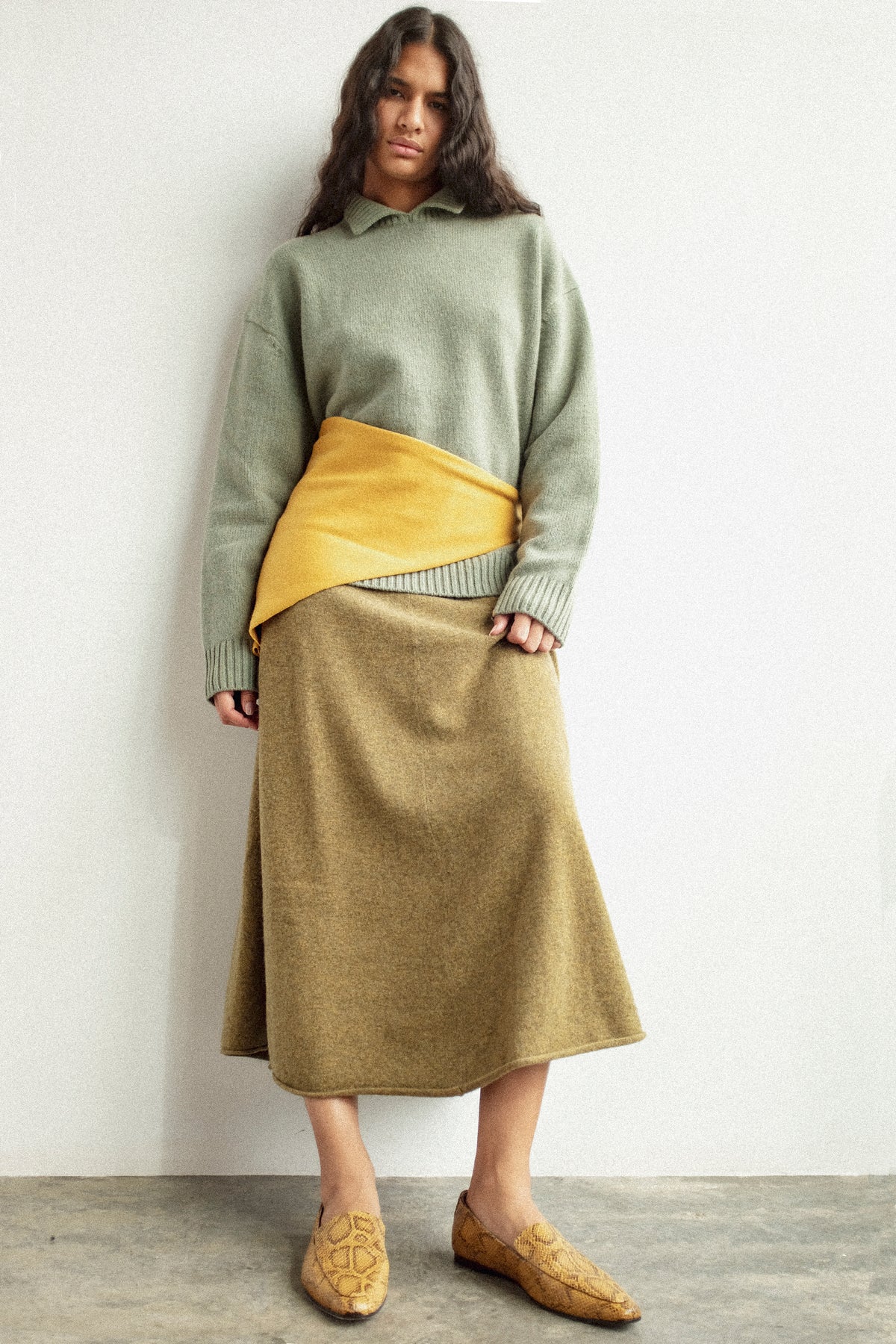 A woman standing against a white wall wearing pieces from Extreme Cashmere's FW26 collection.