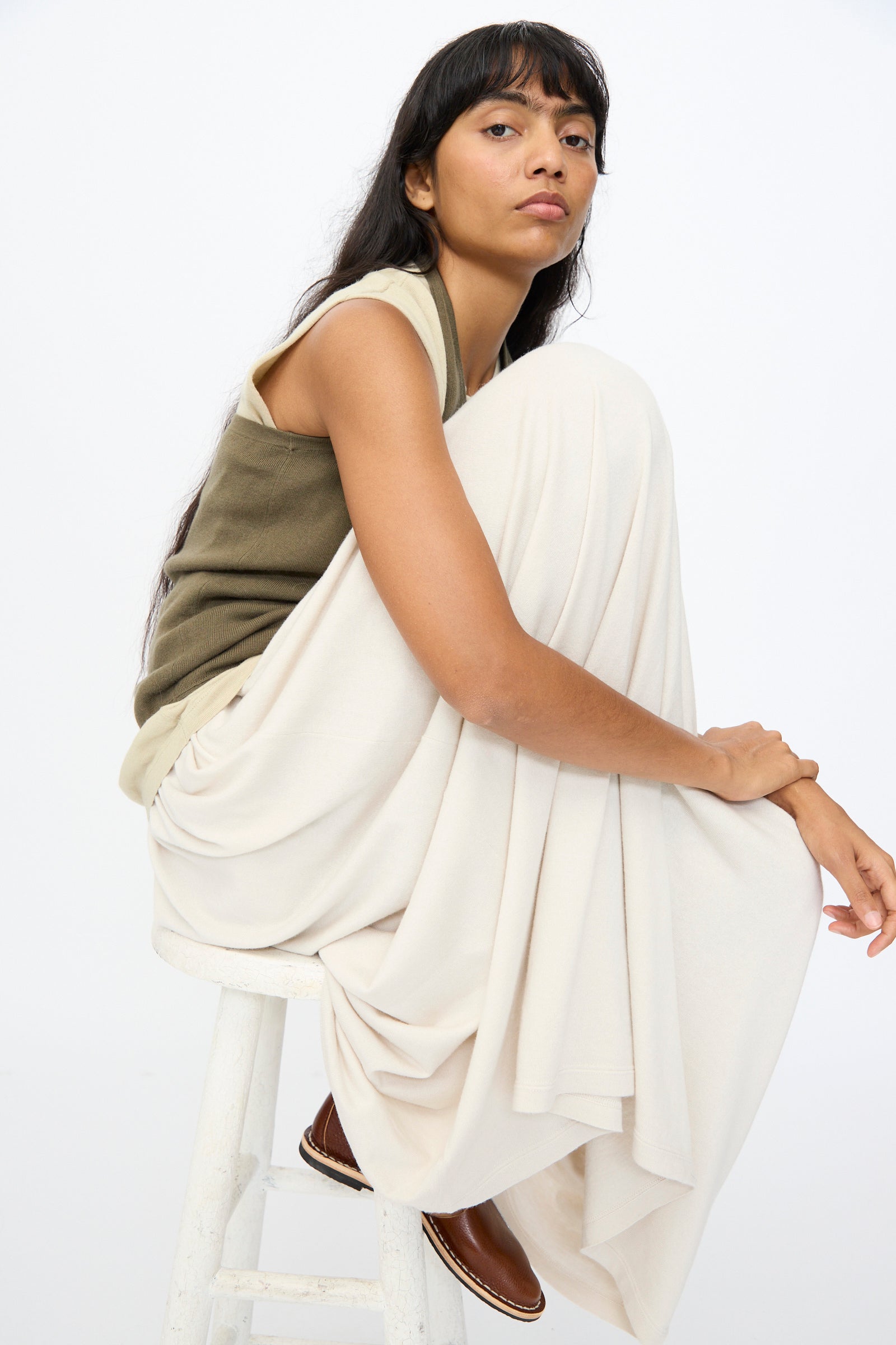 A woman with long dark hair sits sideways on a white stool, wearing the Extreme Cashmere No. 409 Jerry Top in Army, a flowing cream skirt, and brown shoes against a plain white background.
