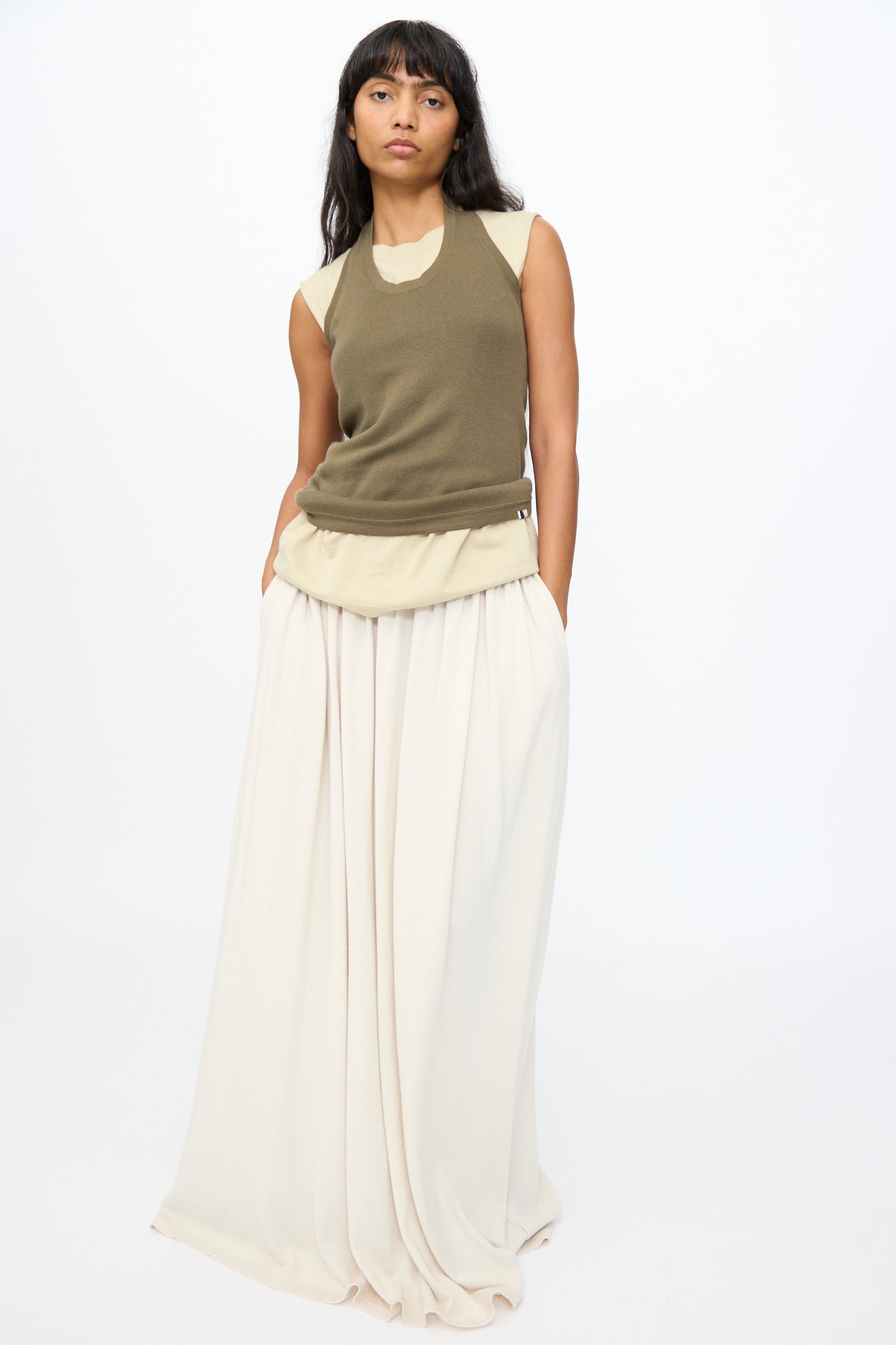 A woman stands against a white background, wearing the Extreme Cashmere No. 409 Jerry Top in Army over a beige shirt and a long cream skirt, with her hands in her pockets.