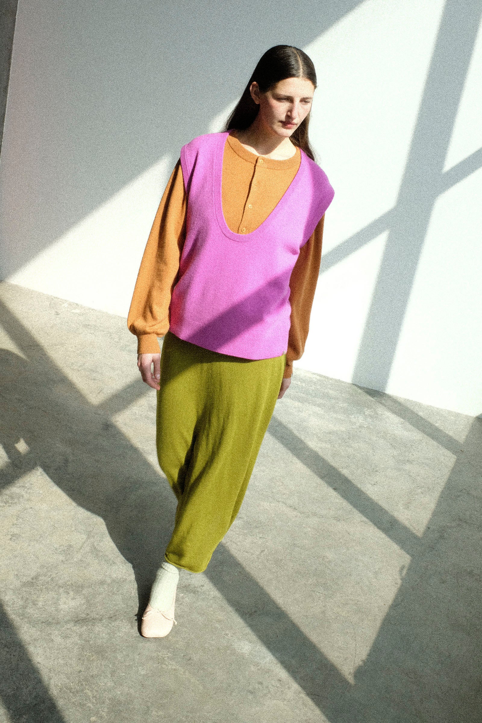 Woman wearing Extreme Cashmere's No. 423 Lord Top in Oleander, No. 280 Bi Sweater in Fox, and the No. 470 Seduce Skirt in Grass, standing on a concrete floor with sunlight casting shadows.