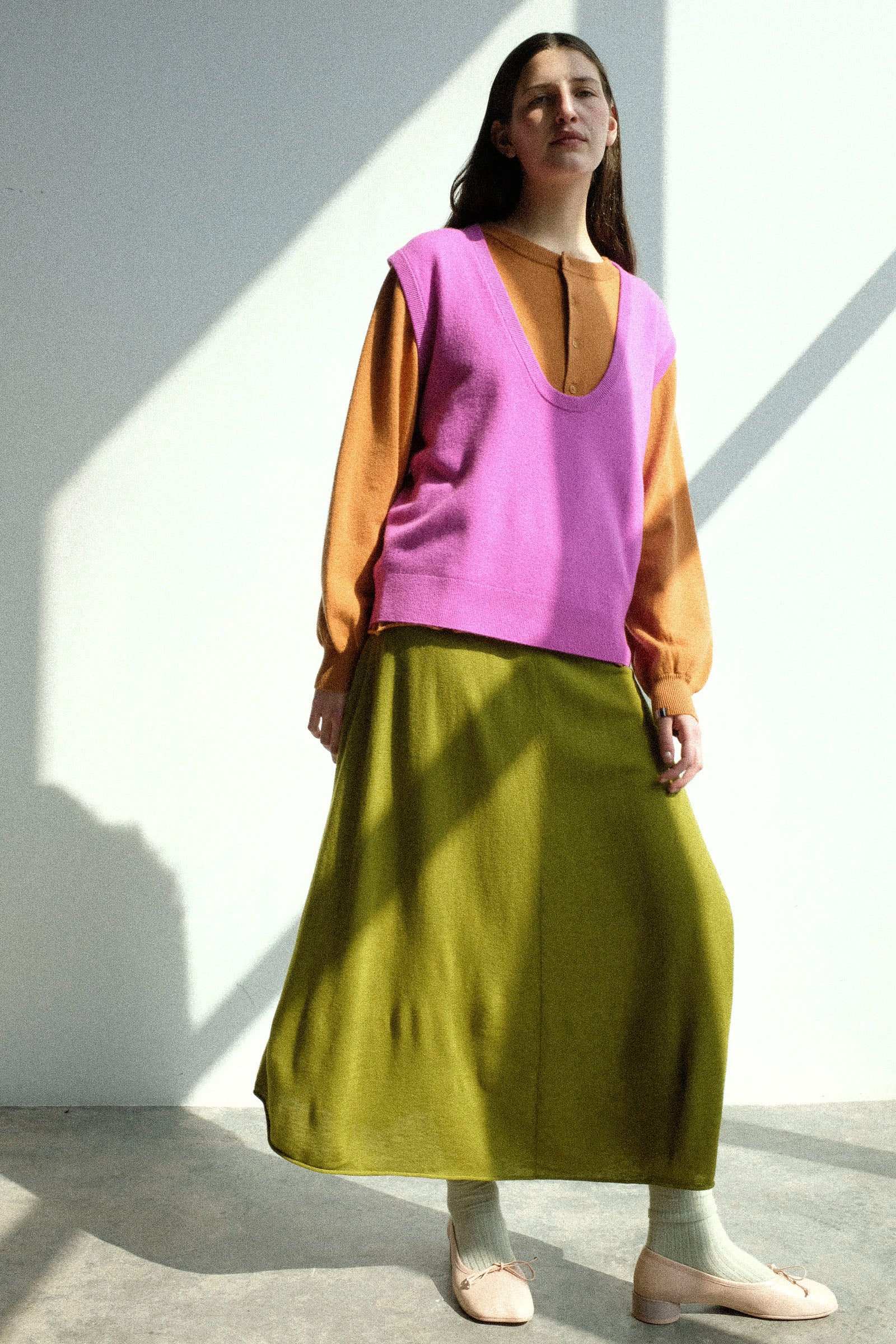 Woman wearing the Extreme Cashmere No. 423 Lord Top in Oleander, No. 280 Bi Sweater in Fox, with the No. 470 Seduce Skirt in Grass, standing against a white wall.