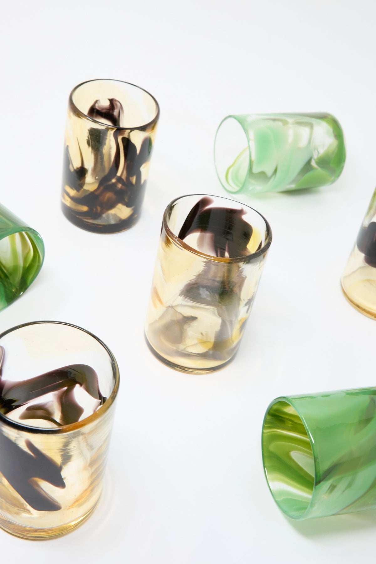 Six Gone Hand Dyed Fumé Glass tumblers arranged on a white surface, featuring swirling patterns in amber, dark brown, and green.