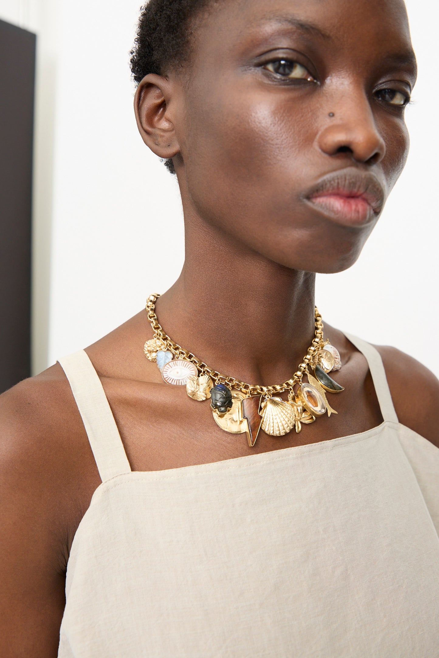 A person in a beige strappy top wears the Grainne Morton Chunky Charm Necklace, handmade in Edinburgh, featuring a chunky 18k gold plated silver chain with assorted sea shell and charm pendants.