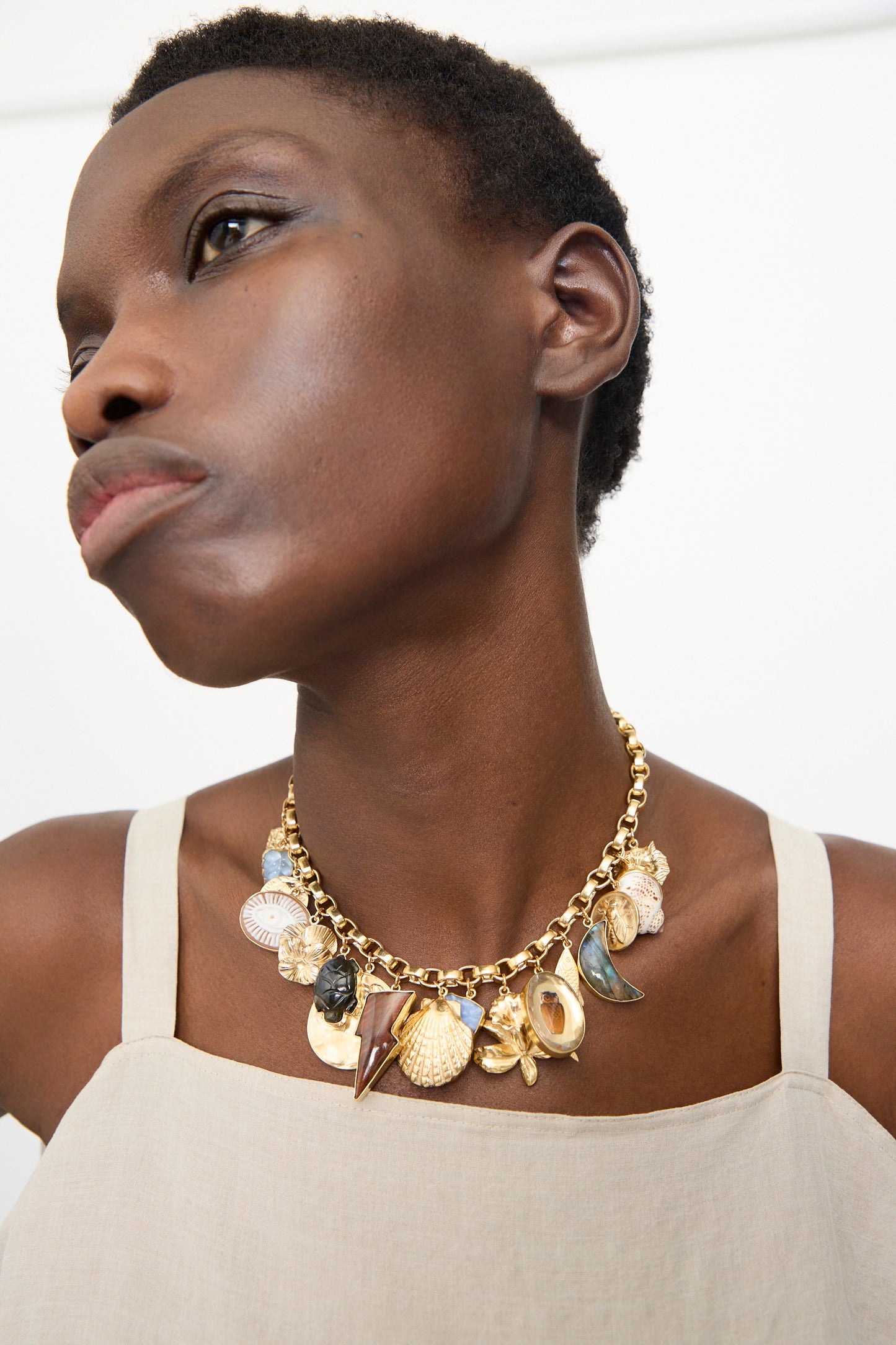 A person wears a beige top and the Grainne Morton Chunky Charm Necklace, a handmade 18k gold plated silver piece from Edinburgh featuring nautical-themed charms like shells.