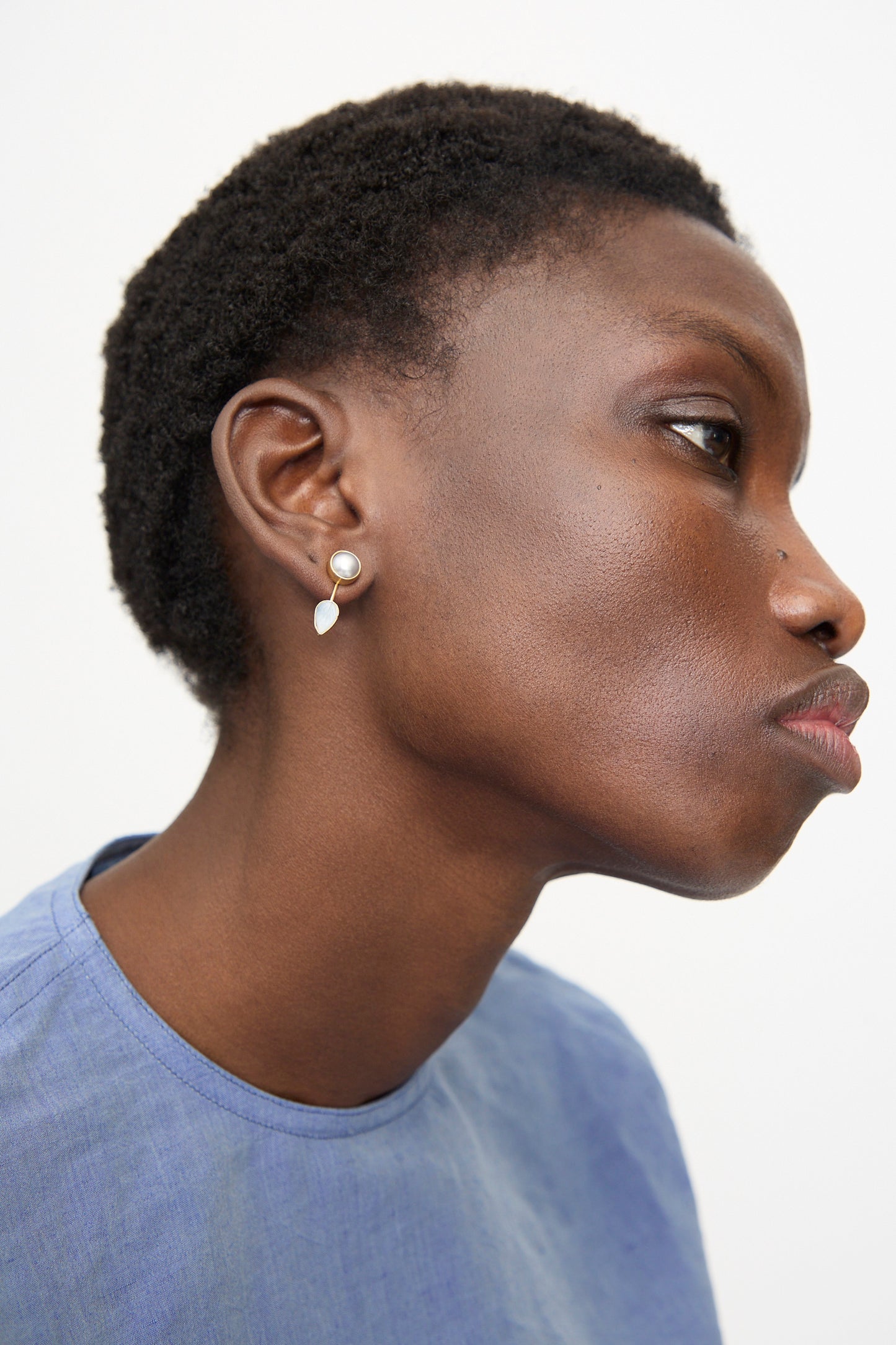 A person with short curly hair in a light blue top wears Grainne Morton’s Detachable Drop Earring in Pearl and Leaf, handmade in 18K gold-plated silver, shown in profile against a plain background.
