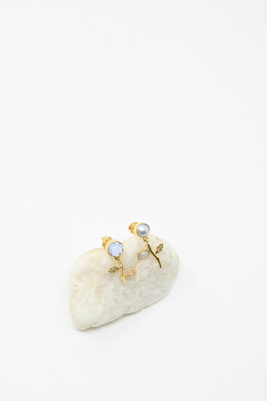 The Grainne Morton Flower Drop Earrings are a mismatched pair, handmade in Edinburgh from 18K gold plated silver with white flower-shaped accents, shown upright on a smooth white stone against a plain background.