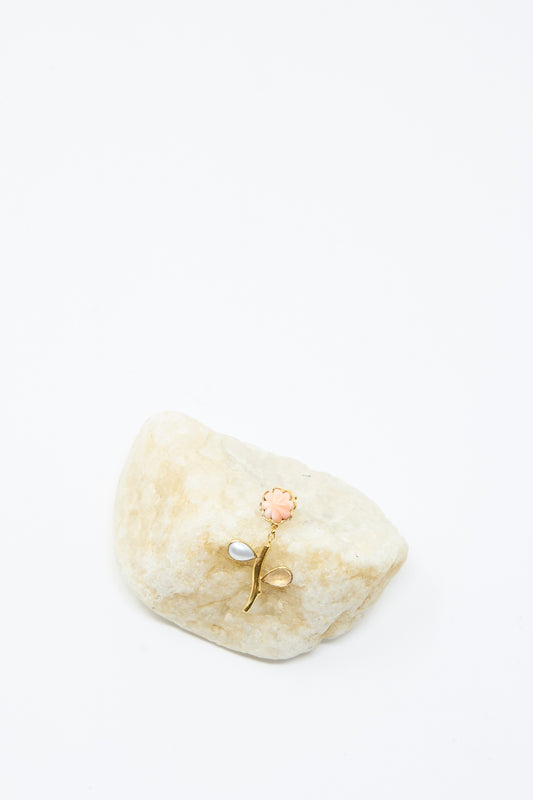 The Grainne Morton Flower Stem Drop Earring, featuring gold petals with pink and white accents, is displayed on a light stone against a white background.
