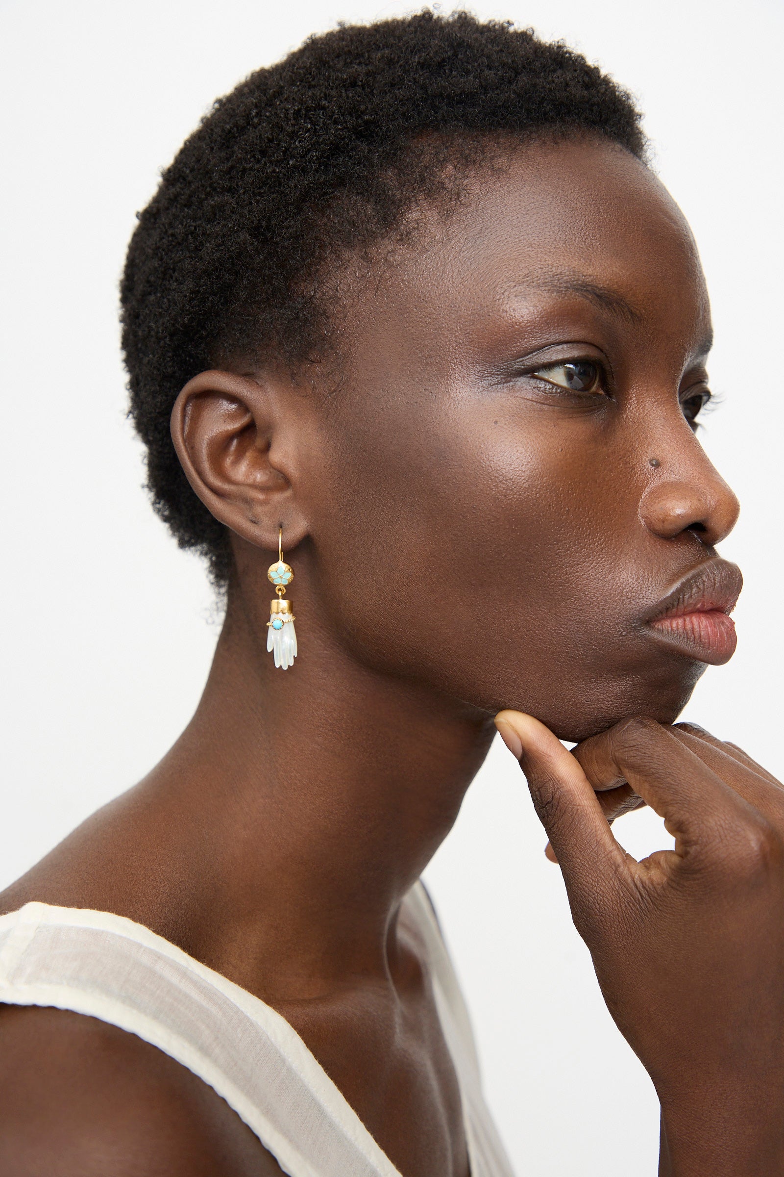A person with short curly hair, in a sleeveless white top, wears Grainne Morton Hand Hooks with Bracelet Earrings (18K gold plated) and rests their chin on their hand while gazing to the side.