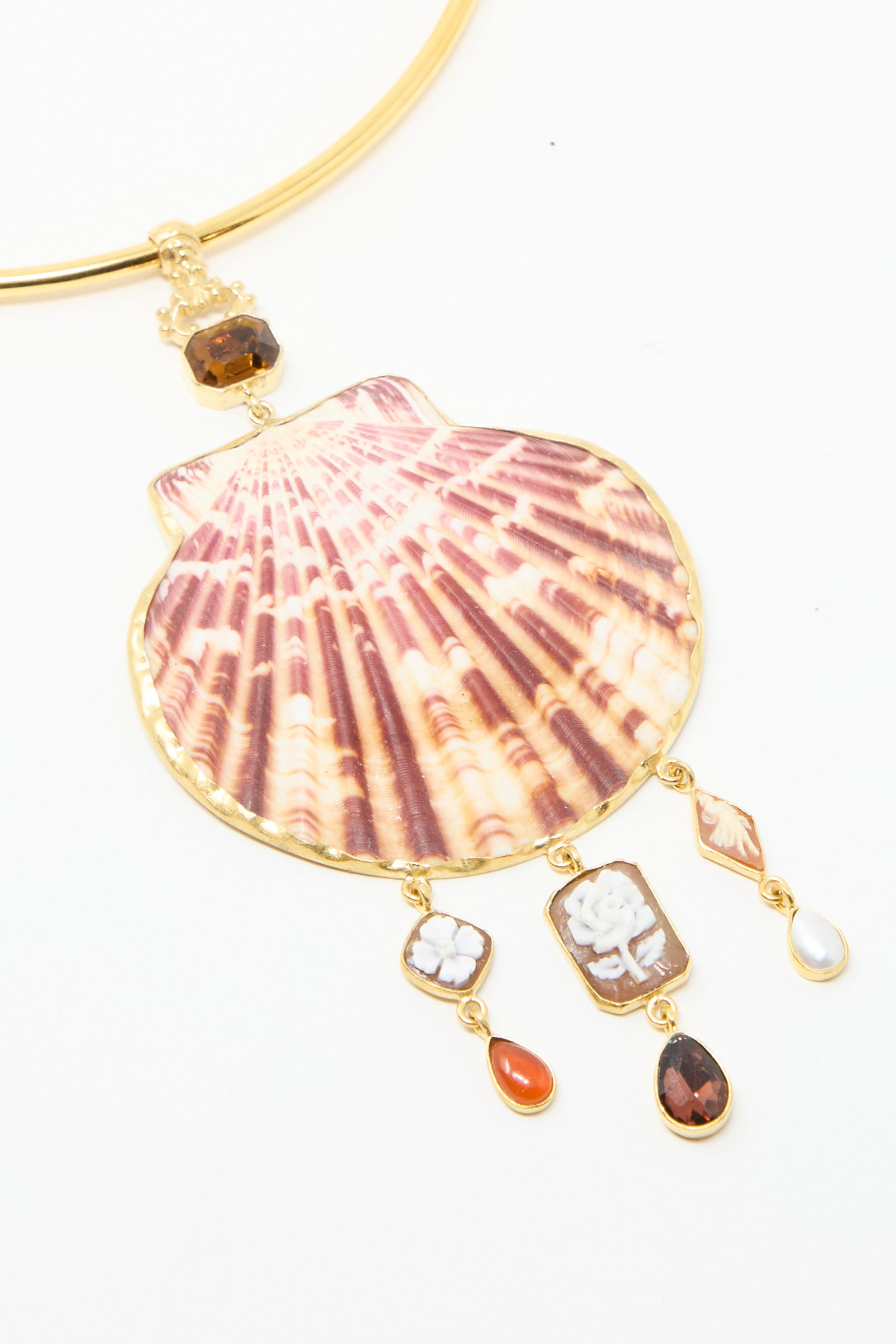 A close-up of the Scallop Necklace by Grainne Morton, a ttorque necklace, featuring a large red striped seashell pendant with hanging charms like gemstones and small cameos, all set in 18k gold-plated silver on a white background.