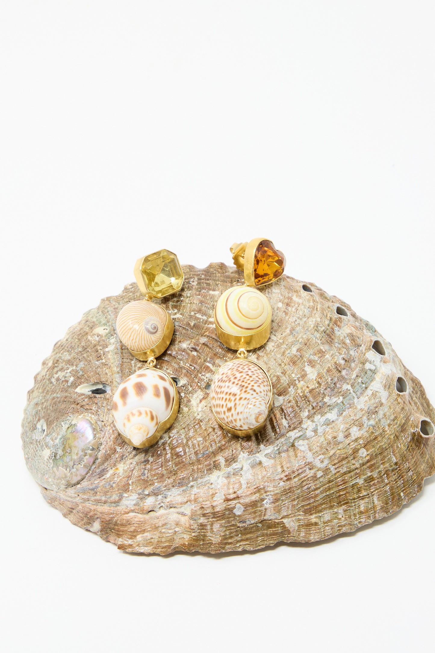 The Shell Charm Drop Earrings by Grainne Morton feature mismatched seashells and amber gemstones, made with 18k gold-plated silver and ethically sourced materials, displayed on a large seashell.