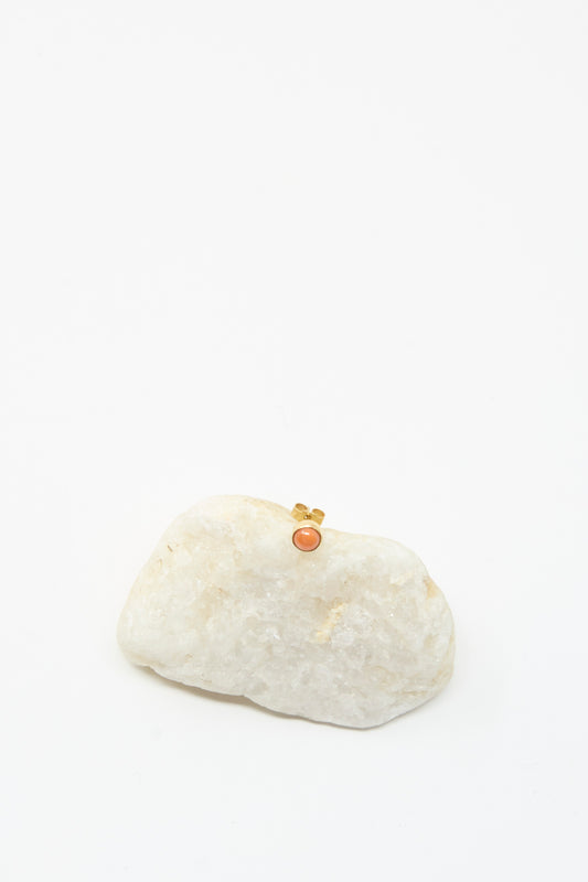 The Stud Earring in Coral by Grainne Morton is showcased atop a white rock against a plain background, its elegance mirrored by the gold-plated ring with an orange stone.