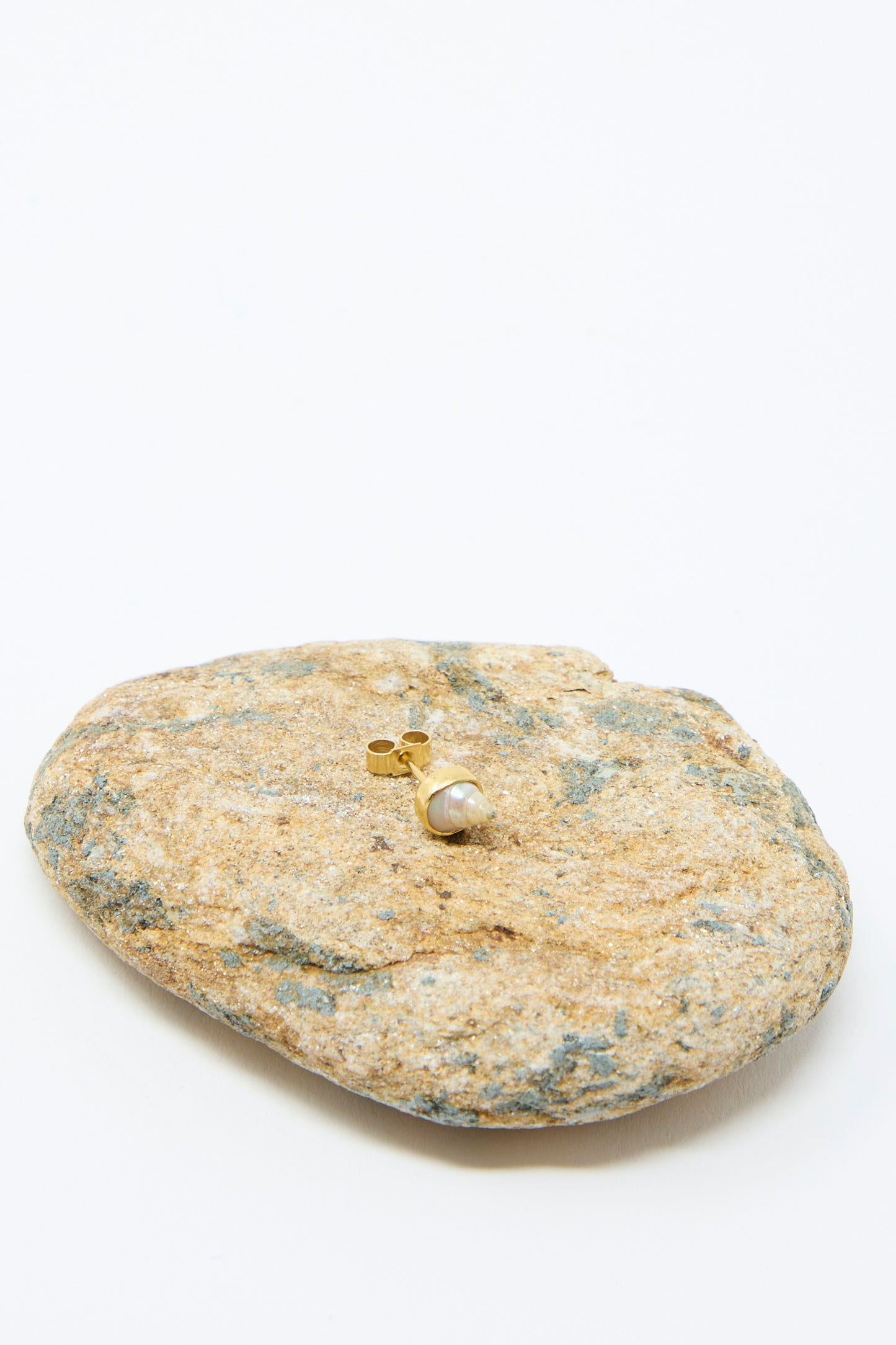 A single "Stud Earring in Shell" by Grainne Morton, made from 18k gold-plated silver with a round pearl, sits on a textured rock against a plain white background.