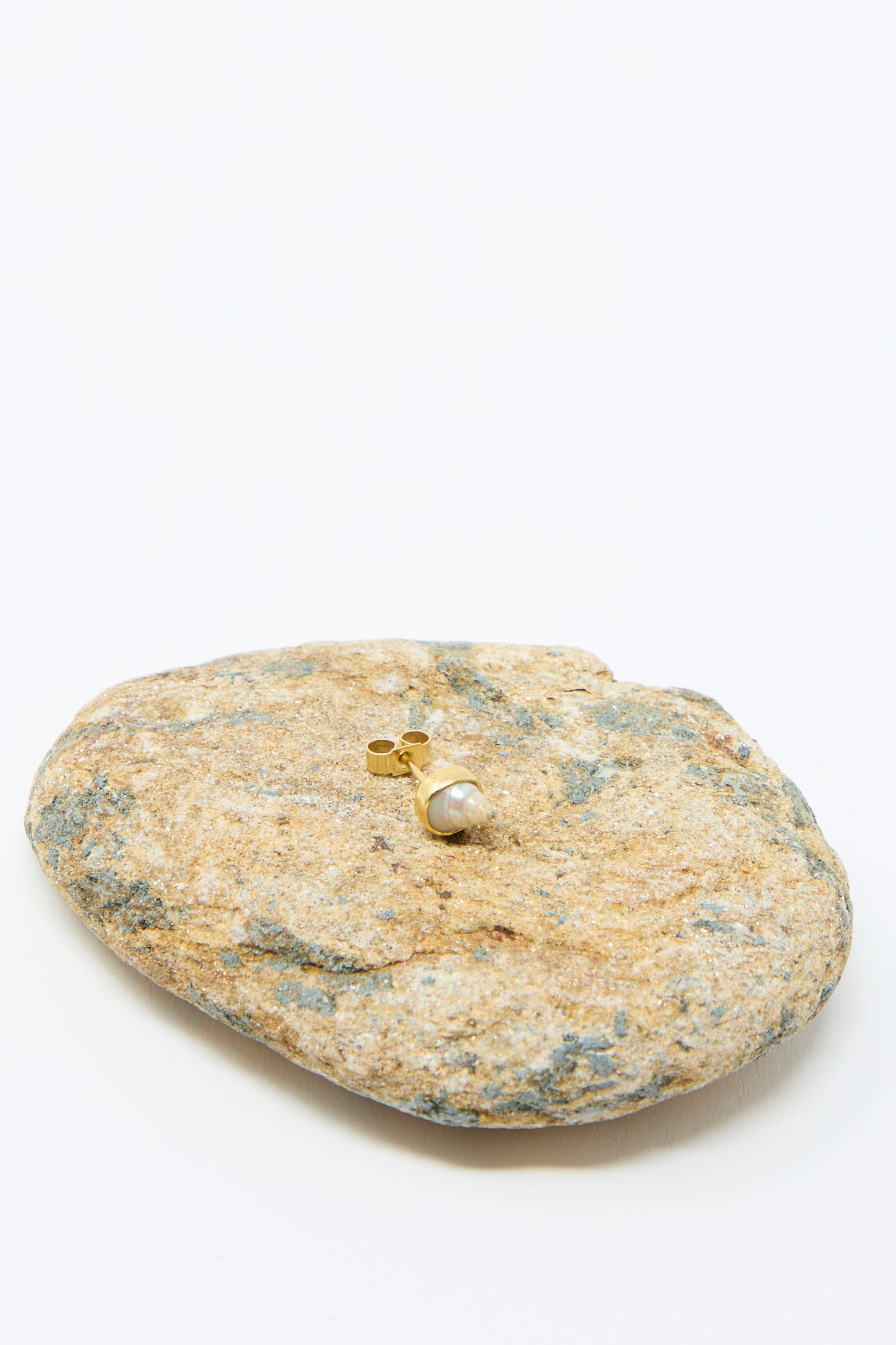 A single "Stud Earring in Shell" by Grainne Morton, made from 18k gold-plated silver with a round pearl, sits on a textured rock against a plain white background.