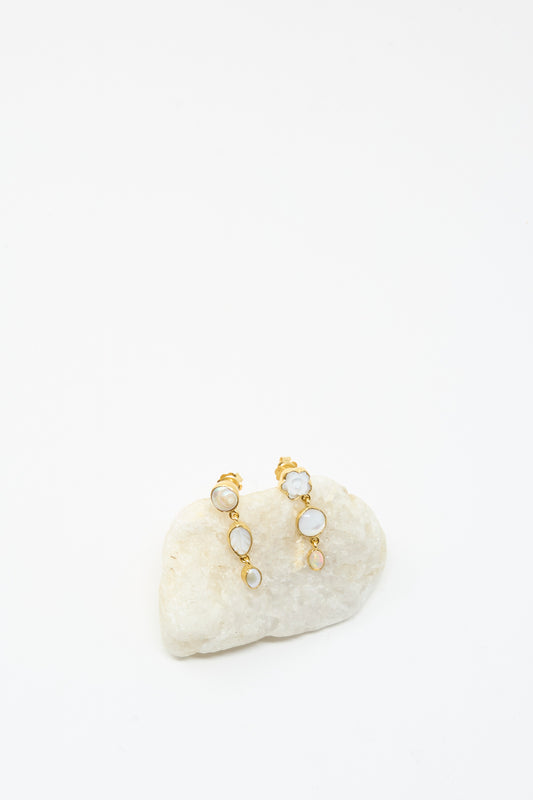 The Grainne Morton Three Charm Moving Drop Earrings, 18K gold plated with circular mother of pearl stones, are displayed on a smooth white rock against a plain white background.