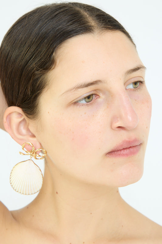 A person wears exquisite Bow and Shell Earrings by Grainne Morton, handcrafted in Edinburgh. These elegant 18k gold-plated silver drop earrings, shaped like shells, beautifully complement their look as they gaze right against a plain backdrop.