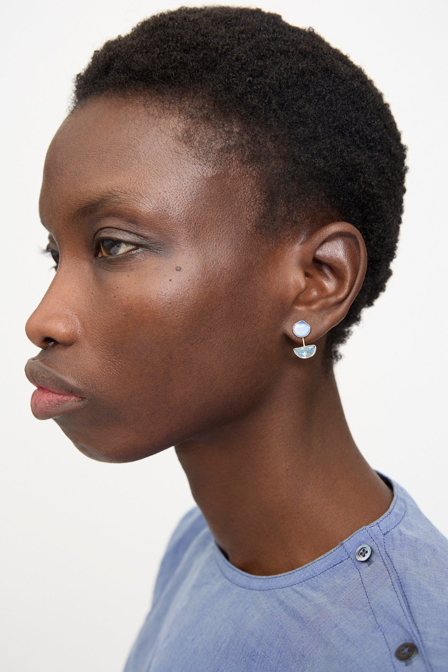 A person with short curly hair wearing a light blue top and the Grainne Morton Detachable Drop Earring in Blue Shell, shown in profile against a plain background.
