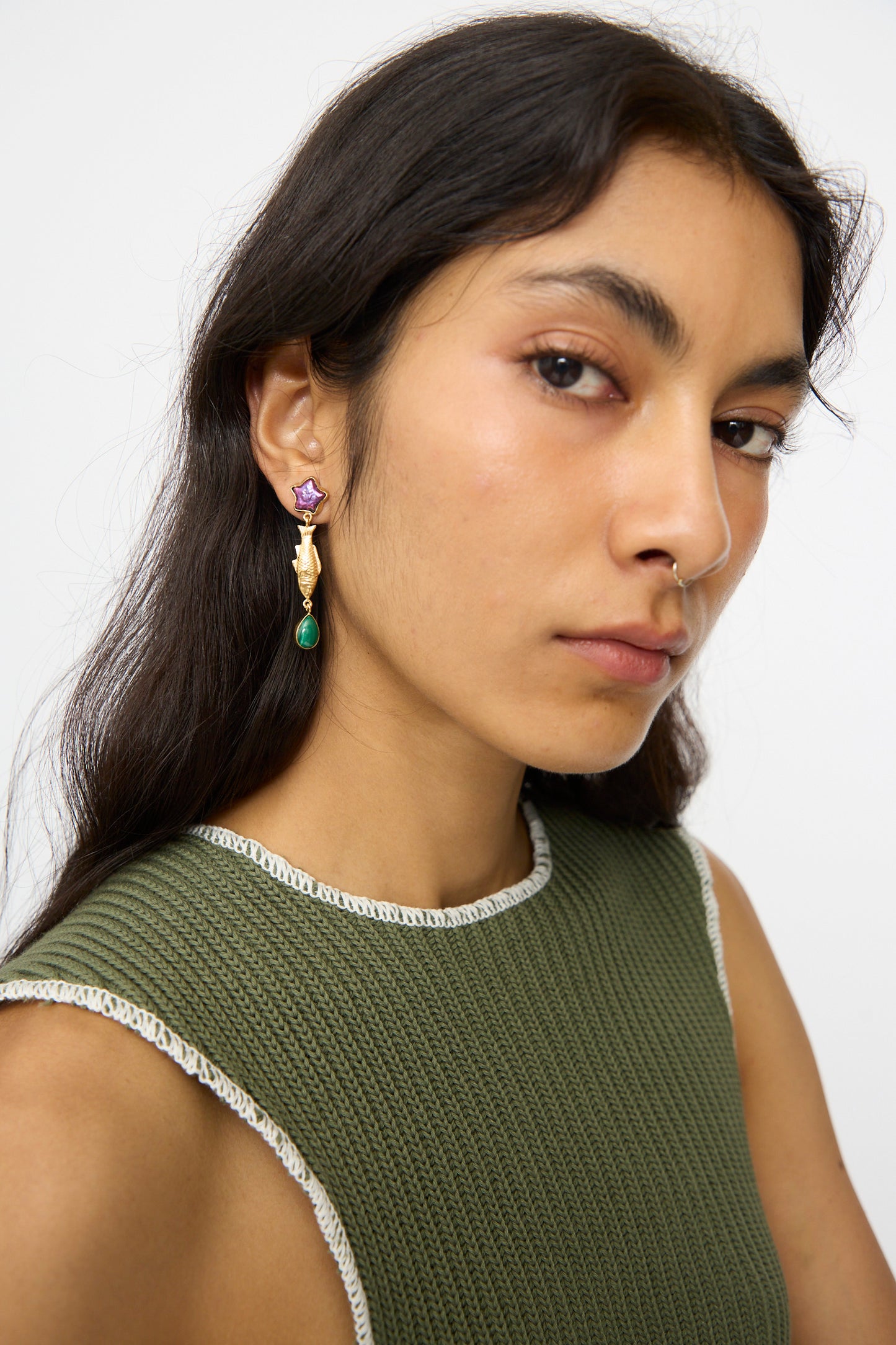 A person with long dark hair wears a textured green sleeveless top and Grainne Morton Fish and Vintage Cabochon Drop Earrings—18K gold plated silver with purple and green details—looking at the camera against a plain background.