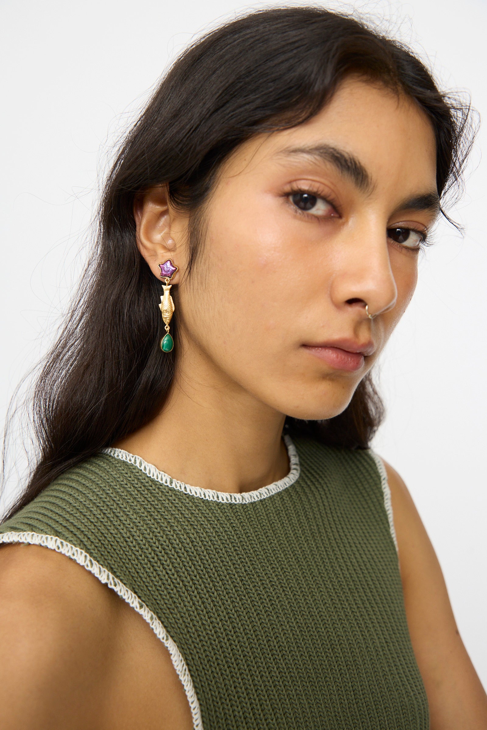 A person with long dark hair wears a textured green sleeveless top and Grainne Morton Fish and Vintage Cabochon Drop Earrings—18K gold plated silver with purple and green details—looking at the camera against a plain background.