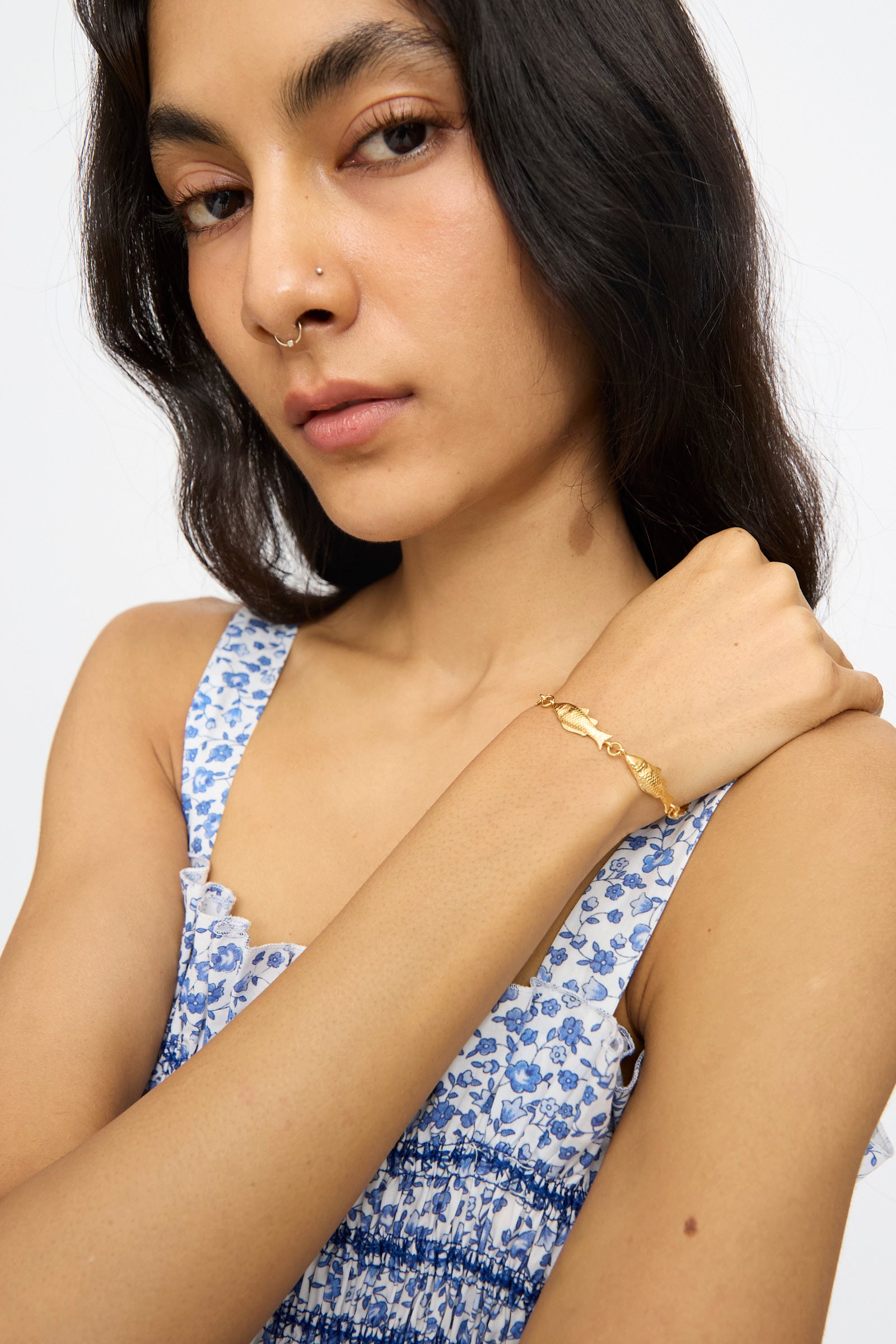 A woman with long dark hair and a nose ring wears a blue and white floral dress and the Grainne Morton Fish Bracelet, posing with her hand near her chin against a plain background.